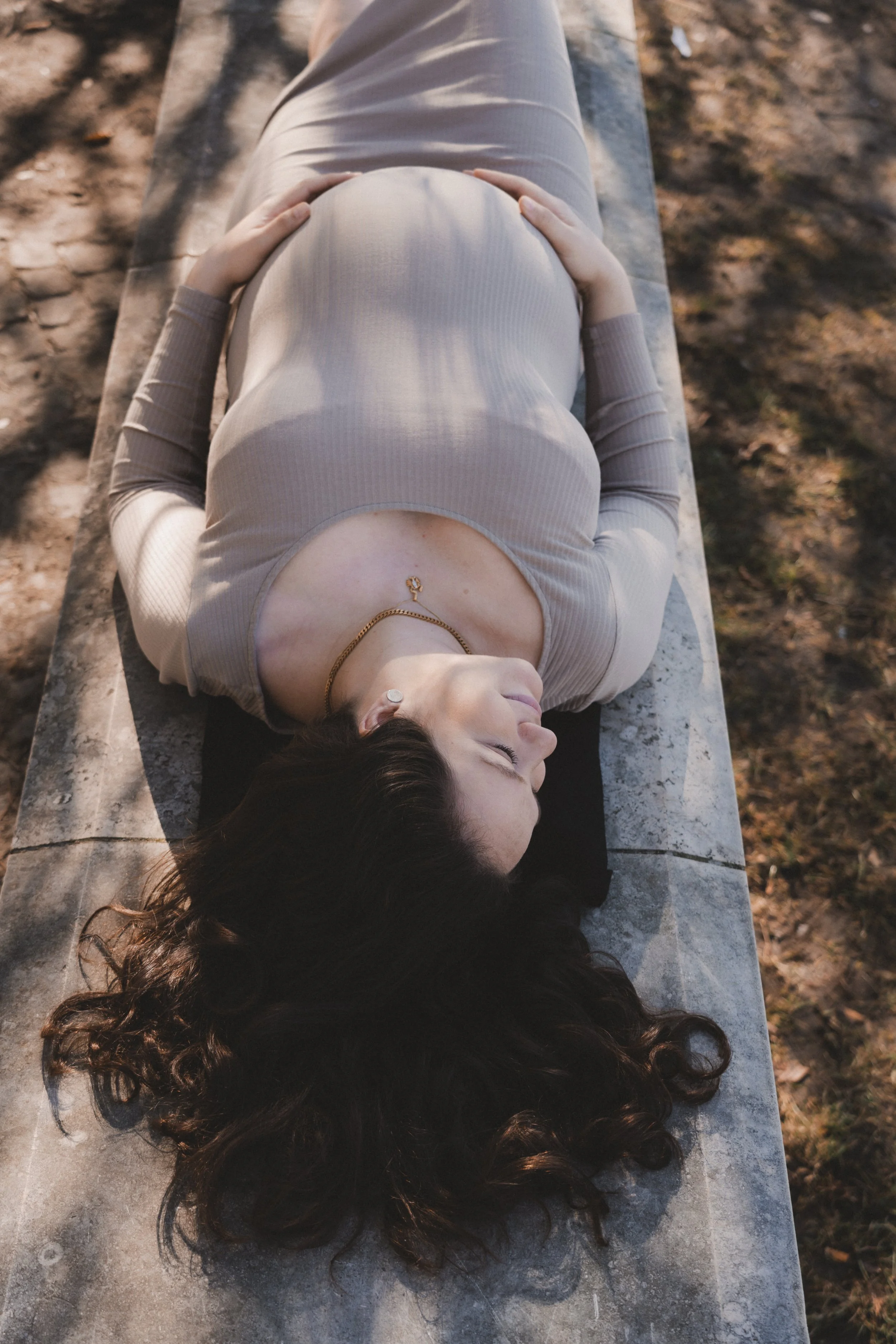 Eine schwangere Frau liegt entspannt auf einem Steinbank im Freien, mit geschlossenen Augen und langen, dunkelbraunen lockigen Haaren.