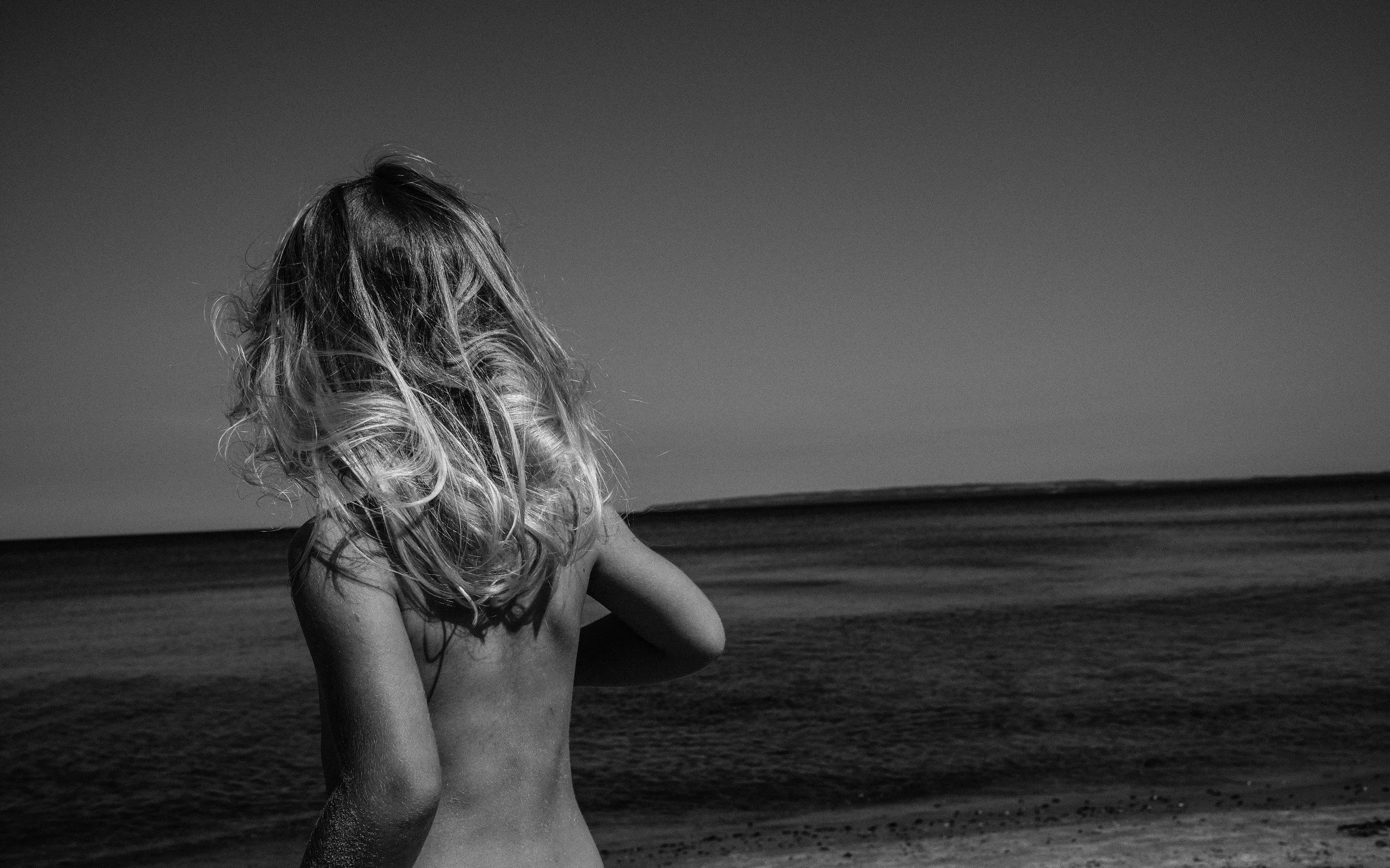 Schwarz-weißes Porträt eines Mädchens am Meer, das in den Wind schaut – poetische Familienfotografie über Freiheit und Kindheit.