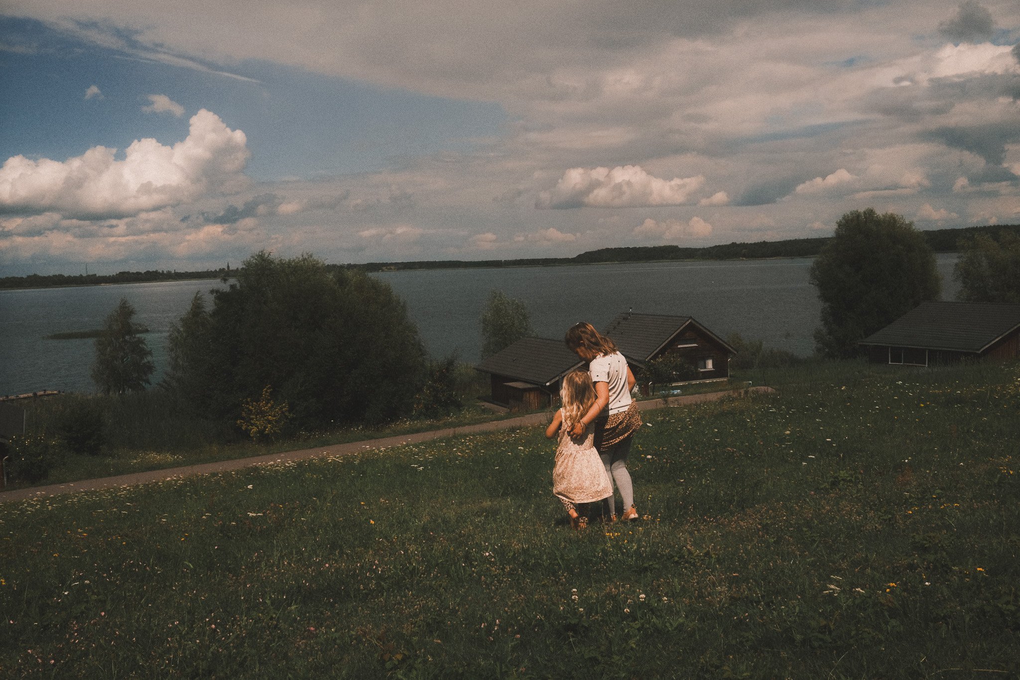 ZZwei Mädchen gehen über eine weite Sommerwiese mit Blick auf einen See – natürliche Kinderfotografie voller Freiheit und Weite.