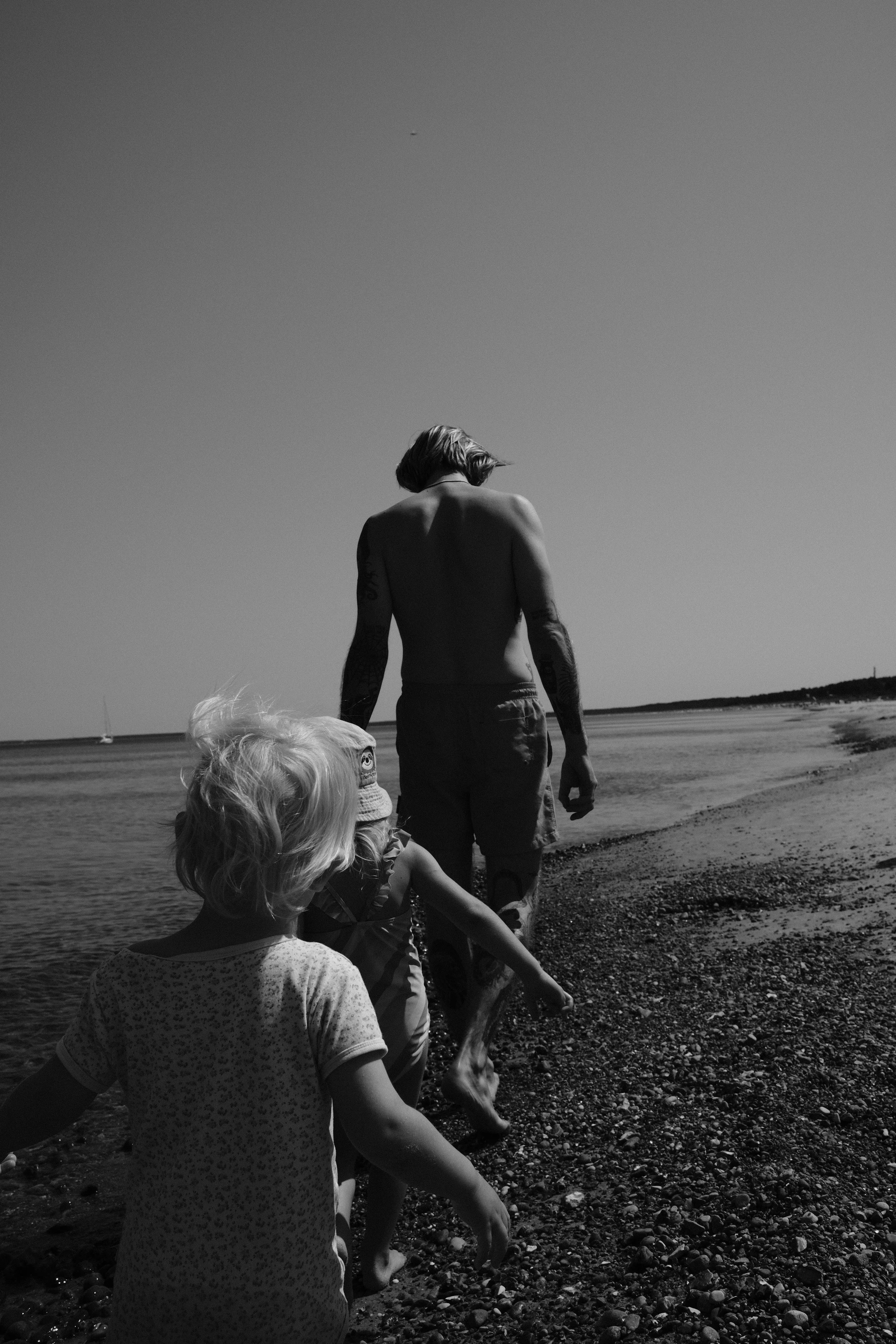 Ein Mann mit Tattoos und kurzen Haaren geht barfuß am Strand, während drei kleine Kinder ihm beim Gehen folgen.