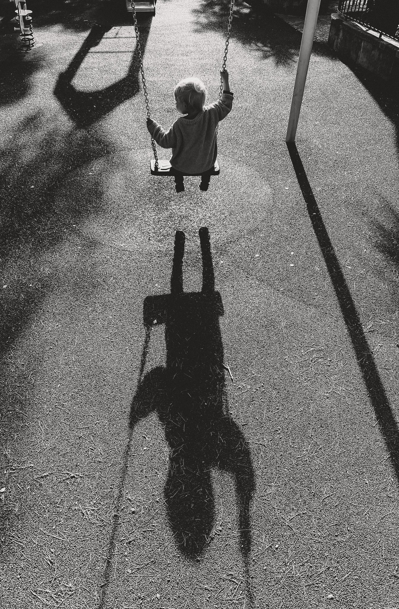 Schwarz-weiß-Foto eines Kindes auf einer Schaukel, dessen Schatten sich auf dem Boden abzeichnet – poetische, zeitlose Kinderfotografie.