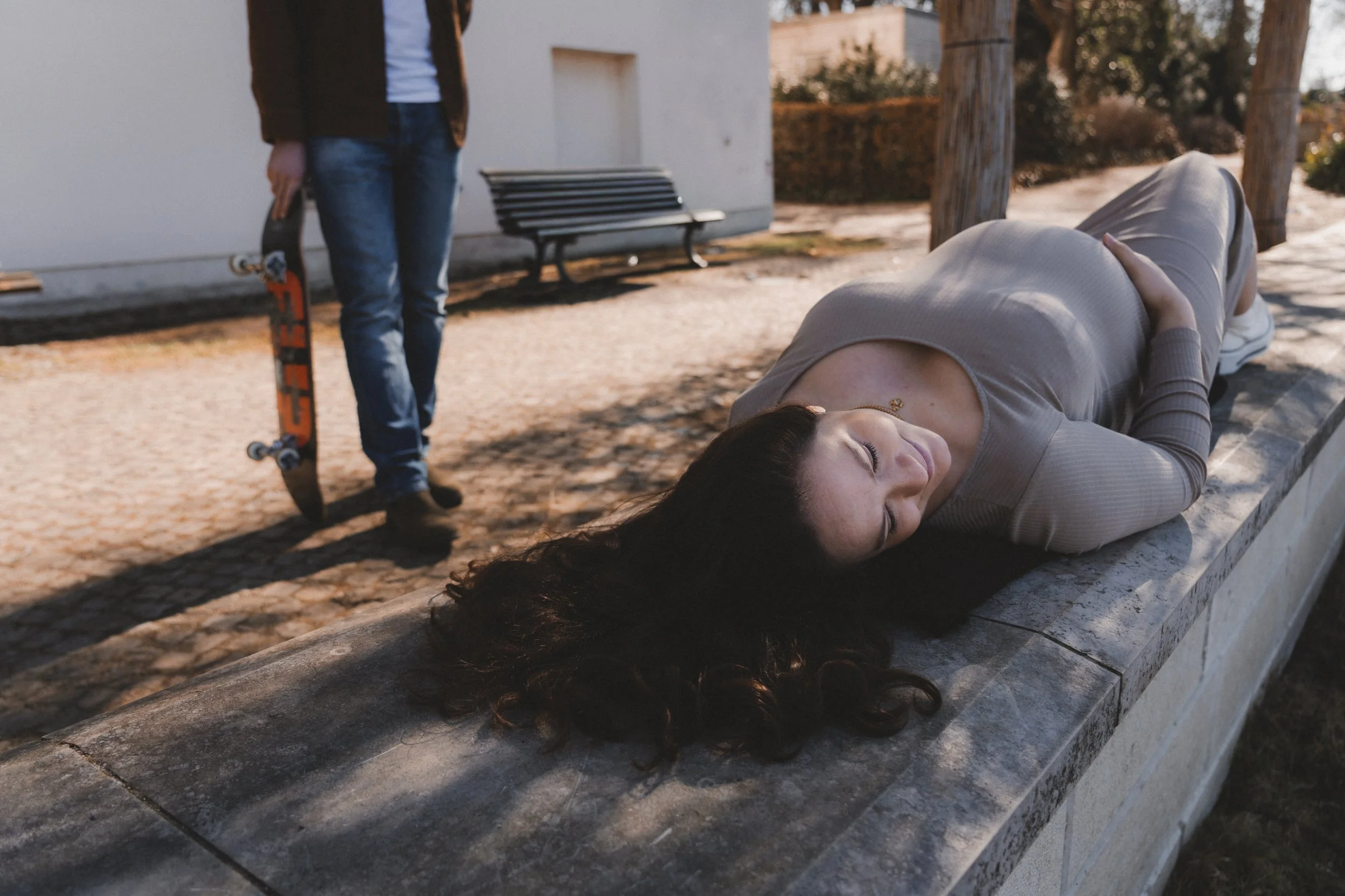 Eine Frau liegt auf einer Steinbank im Freien, scheint zu schlafen oder bewusstlos zu sein, während ein junger Mann mit einem Skateboard in der Hand in der Nähe steht.
