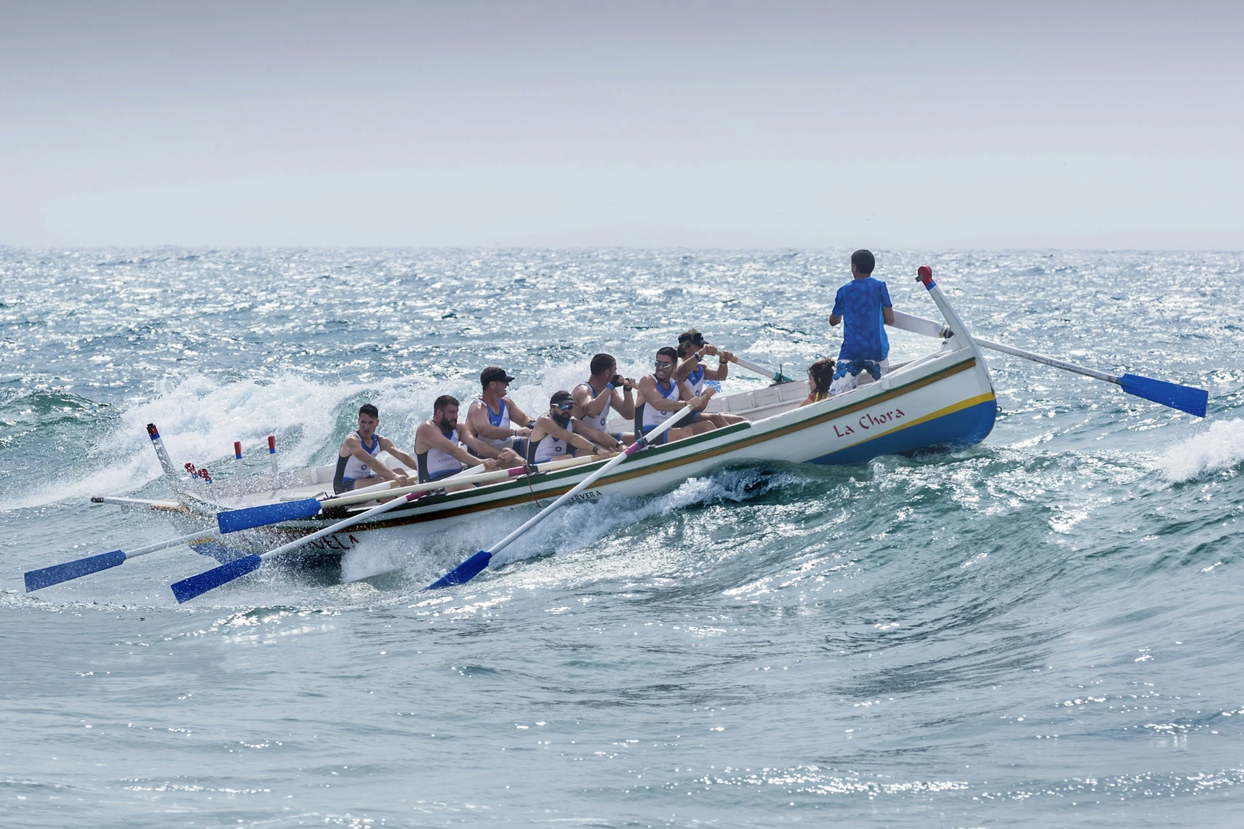 Eine Gruppe von Menschen beim Ruderbootrennen auf dem Meer, alle tragen sportliche Kleidung und Sonnenbrillen, während das Boot namens 'La Chora' durch die Wellen fährt.