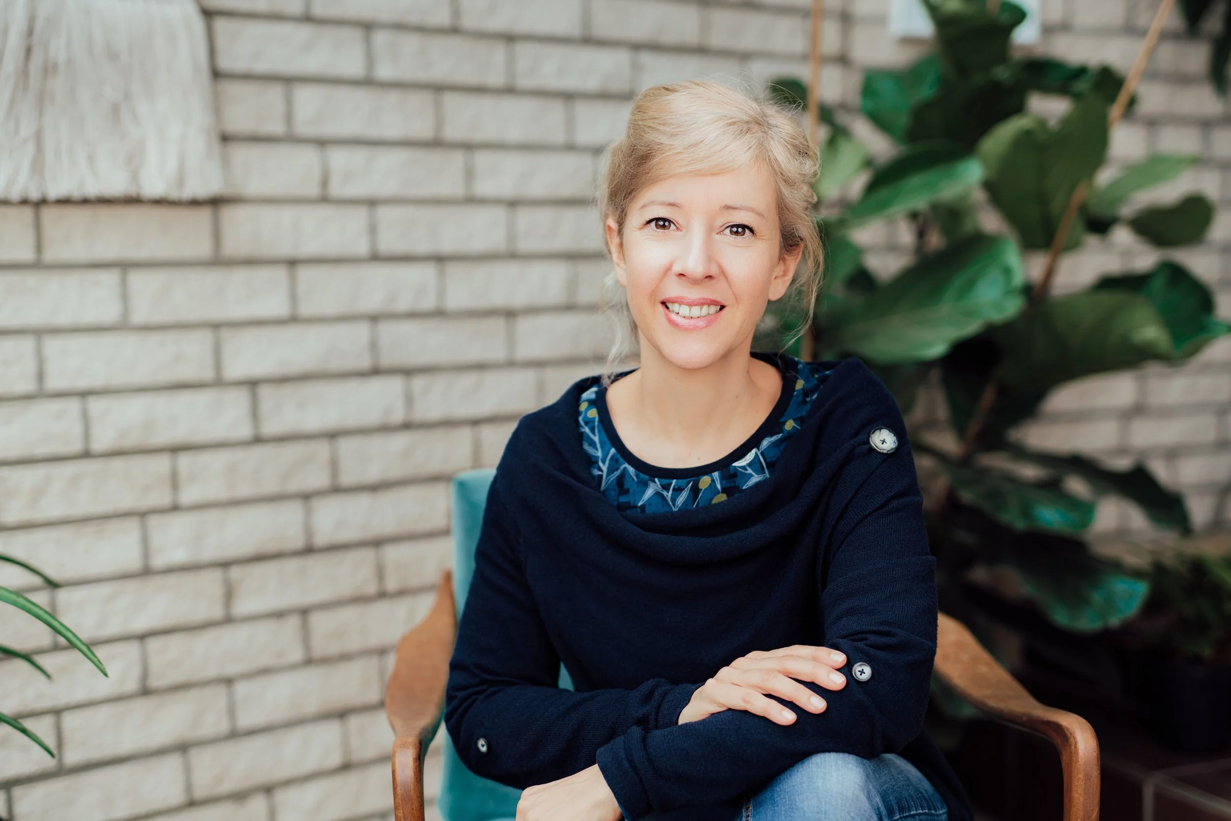 Lächelnde blonde Frau sitzt in einem natürlichen Umfeld vor einer Ziegelwand und grünen Pflanzen.
