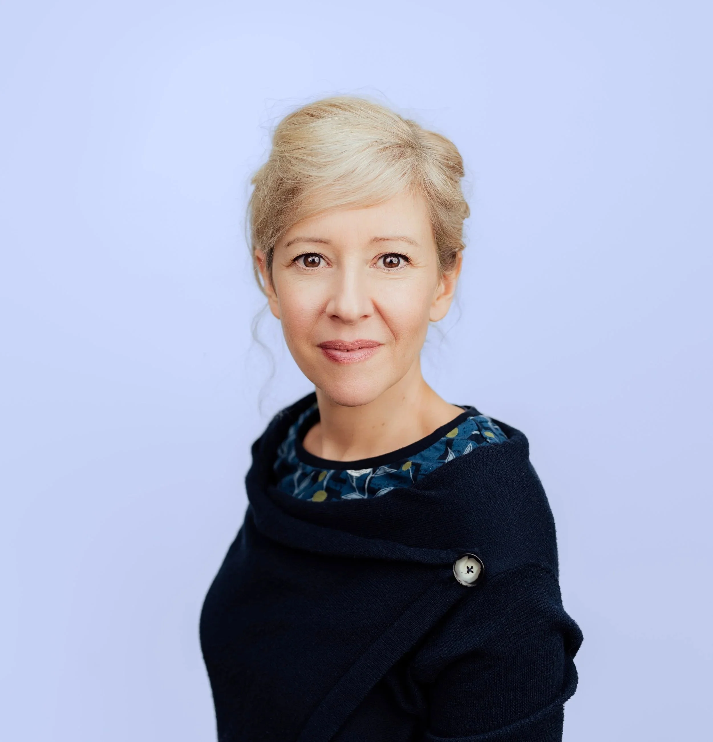 Porträt einer Frau mit blonden Haaren vor einem hellblauen Hintergrund, trägt einen dunklen Pullover mit einem blauen Muster