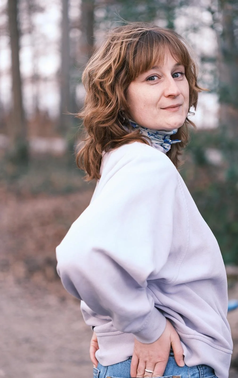 Junge Frau mit kastanienbraunen, lockigen Haaren steht im Freien, trägt einen grauen Pullover und einen blauen Schal, blickt lächelnd zur Seite, im Hintergrund ein Wald.