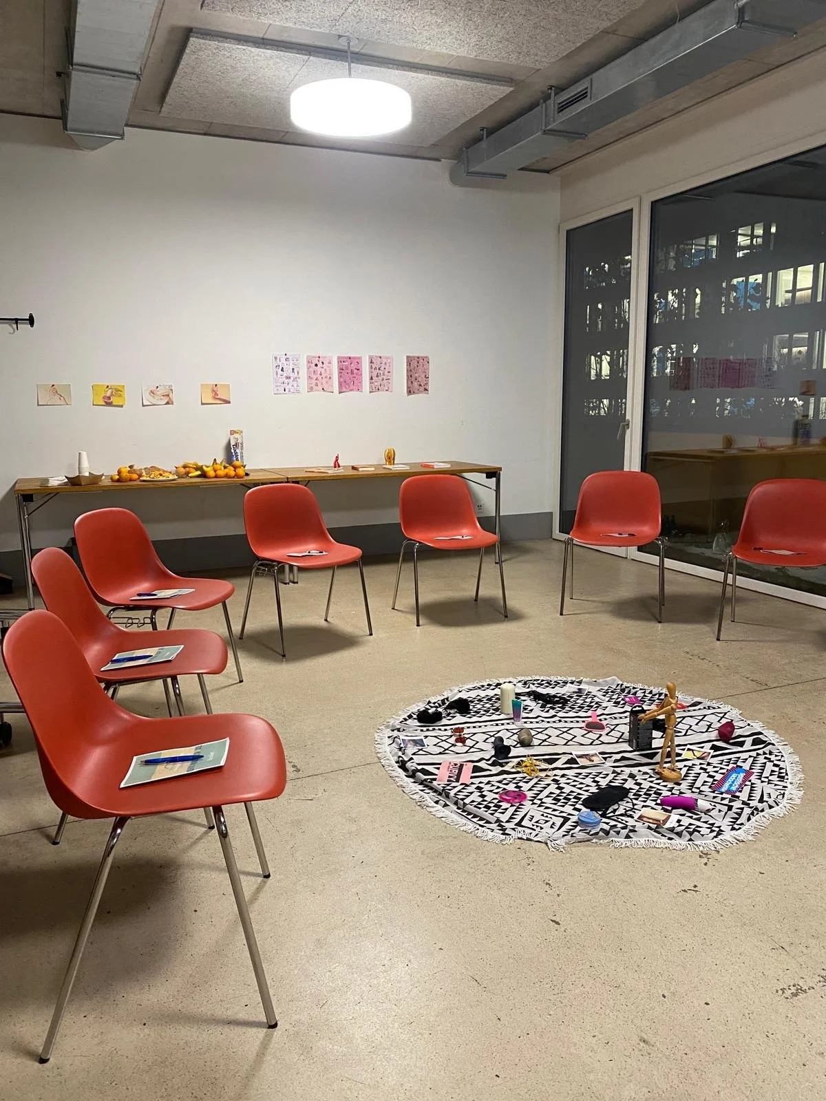 Ein heller Raum mit roten Stühlen in einem Kreis, einem runden Teppich mit Spielzeug in der Mitte, einer längeren Tafel an der Wand mit Obst und Papieren, und einem Fenster, das auf ein Winterbild zeigt.