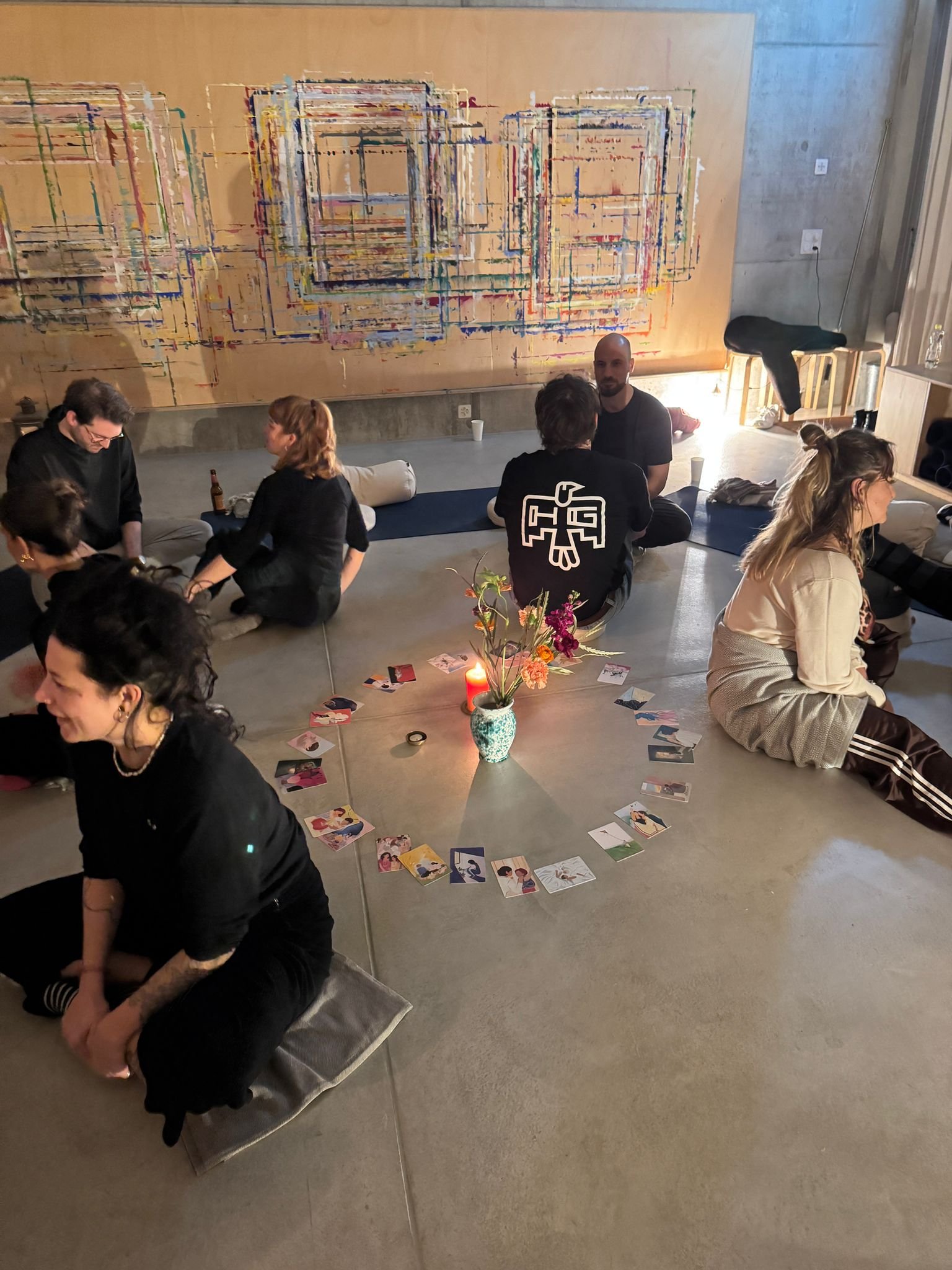 Gruppierung von Menschen bei einem gemütlichen Treffen in einem Raum mit abstrakter Wandkunst, in der Mitte ein Blumenarrangement und Karten auf dem Boden, warme Beleuchtung.