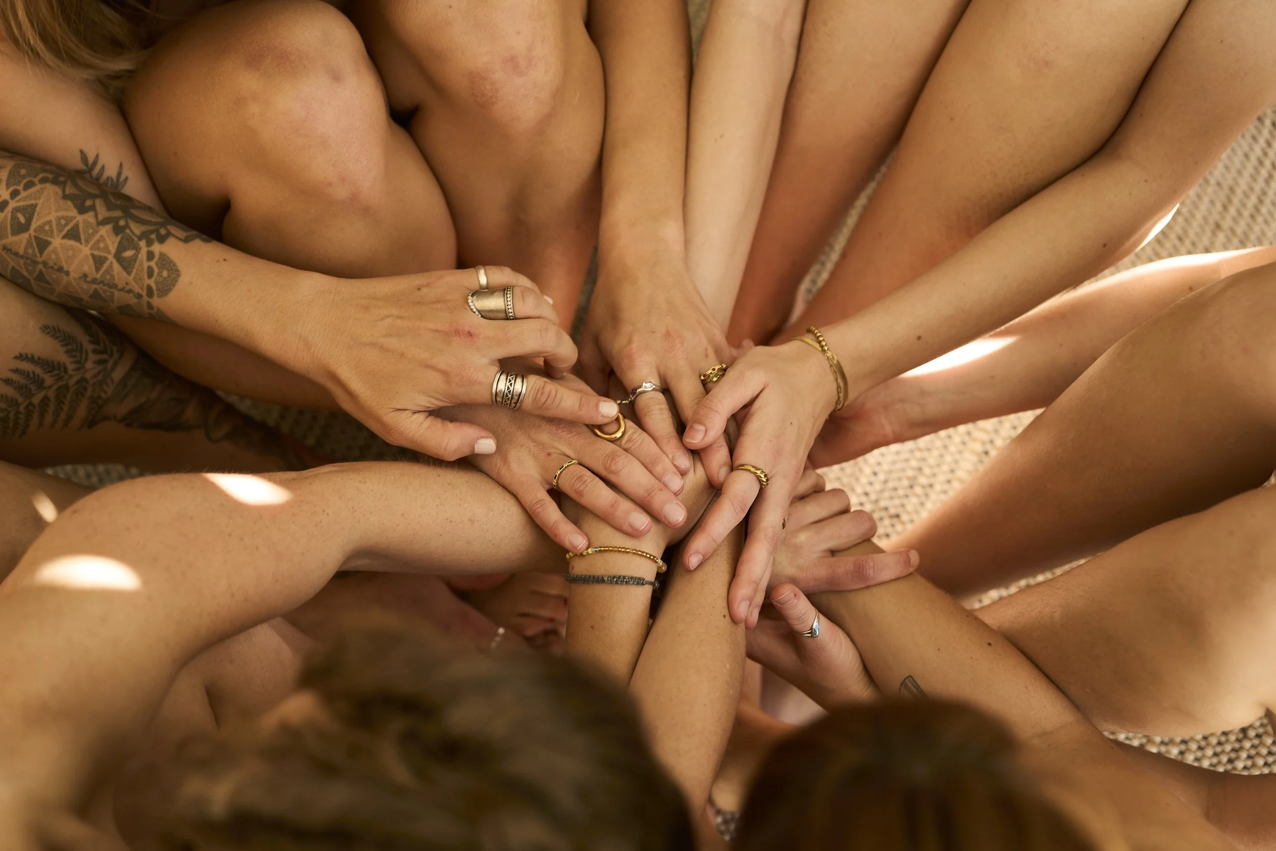 Mehrere nackte Menschen legen ihre Hände übereinander, sichtbar mit verschiedenen Ringen, auf einem kuscheligen, hellen Teppich.