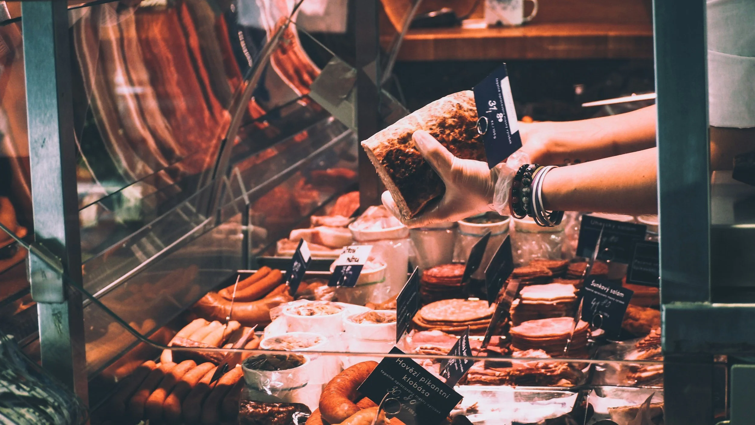 A person in a glove is holding a large piece of meat in a refrigerated display case at a butcher shop or deli. Various cuts of meat and sausages are visible on the shelves below.