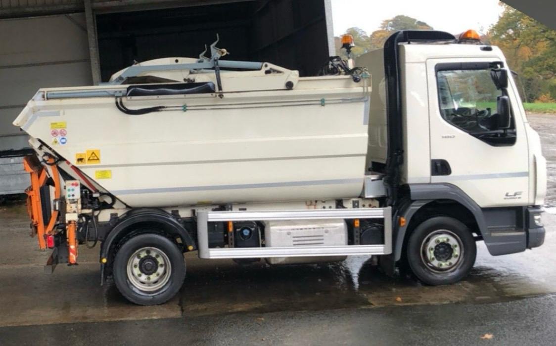 Side view of a street cleaning truck parked on a wet road, with a spray nozzle at the rear and equipment on top.