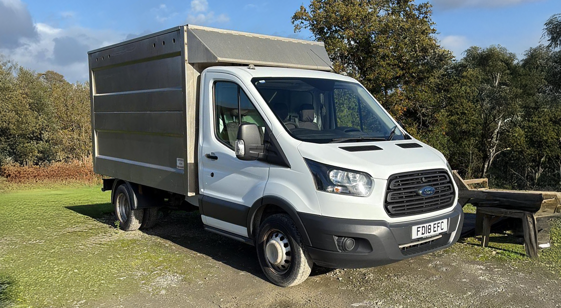 White Ford Transit cargo van with a large enclosed cargo box parked outdoors on grass and gravel with trees and blue sky in the background.
