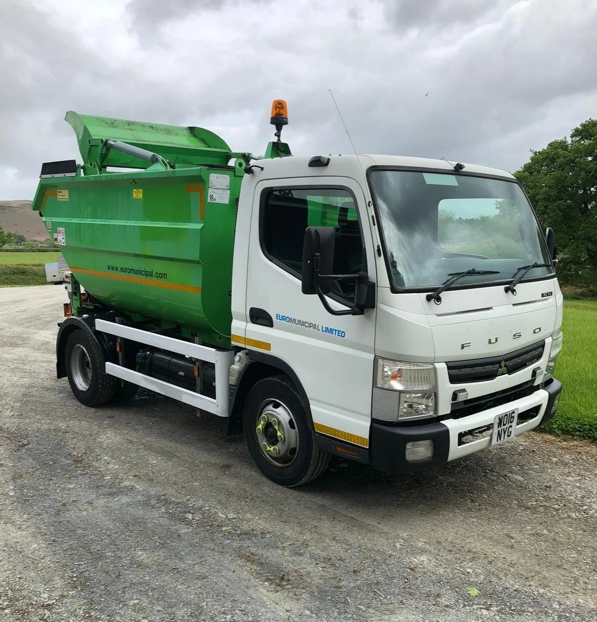 A street cleaning truck painted white and green, with a large green container on the back, parked on a gravel surface near greenery under a cloudy sky.