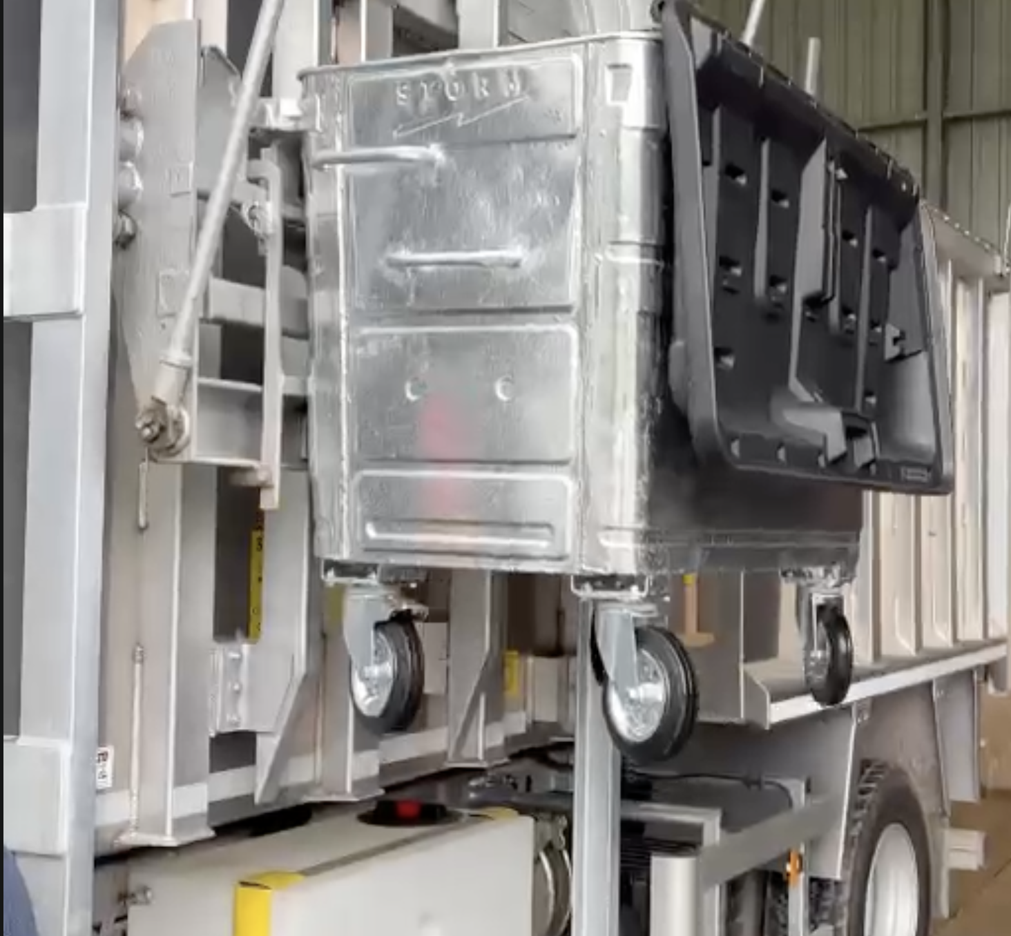 A stainless steel food cart with a black plastic container on the side, situated in an industrial or storage environment.