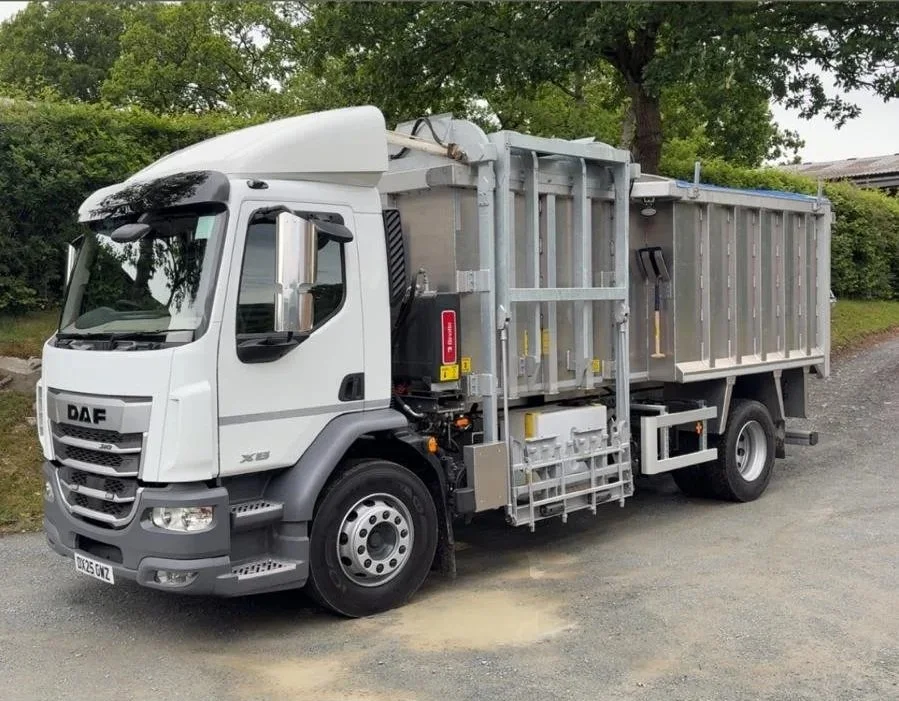 A white DAF flatbed truck with a metal cage over the cargo area is parked on a gravel driveway, with green trees and bushes in the background.