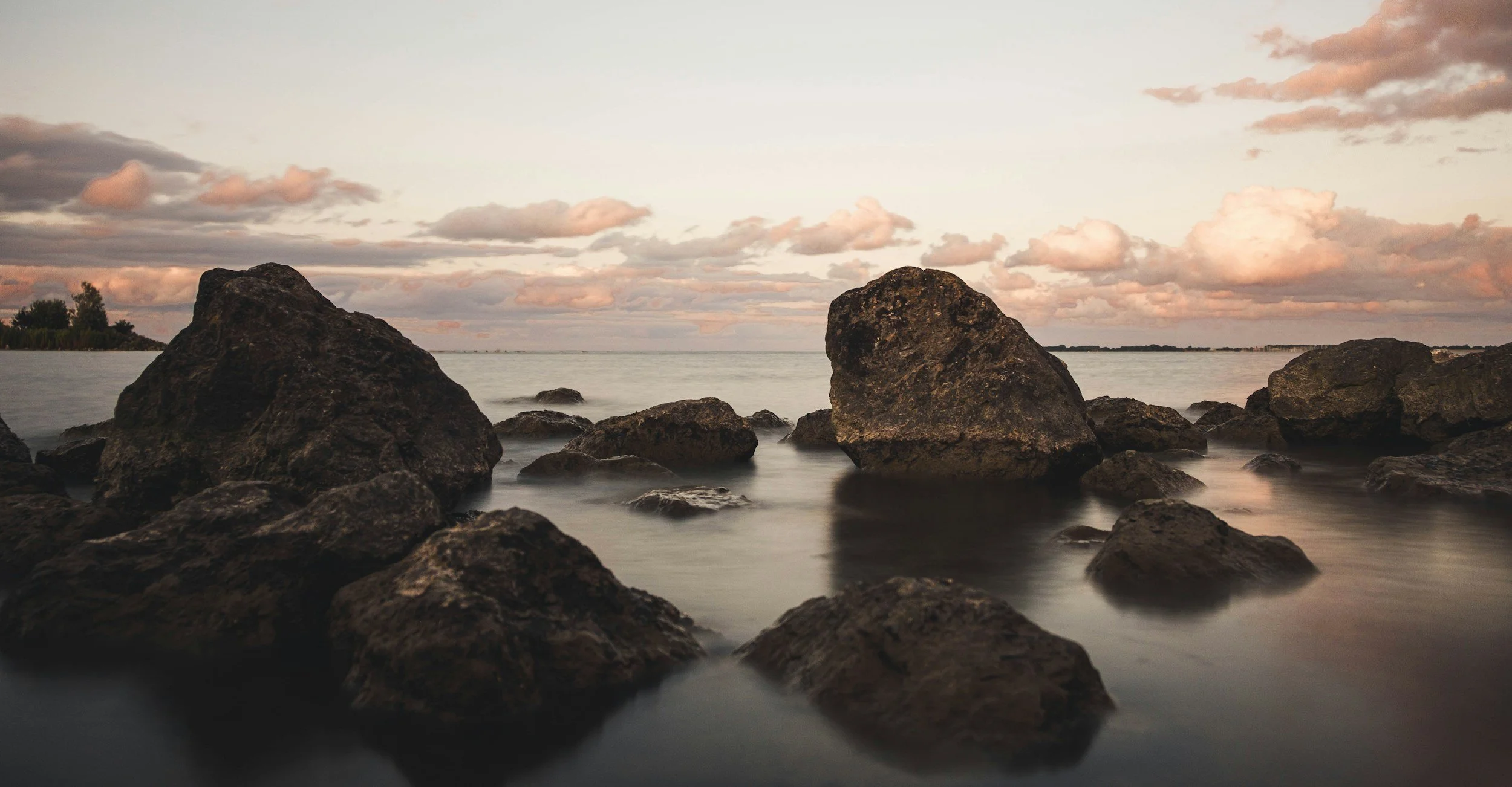 Rocks along shoreline at sunset with cloudy sky reflected in calm water.