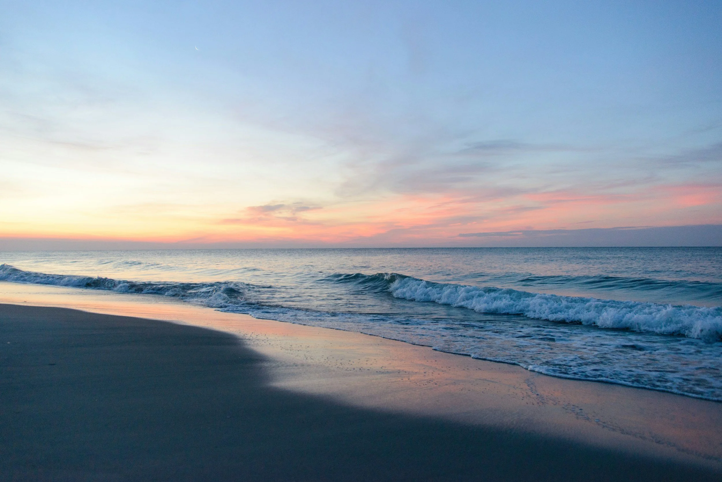 Beach scene at sunset with waves crashing onto the shore, pastel sky with orange, pink, and blue hues.