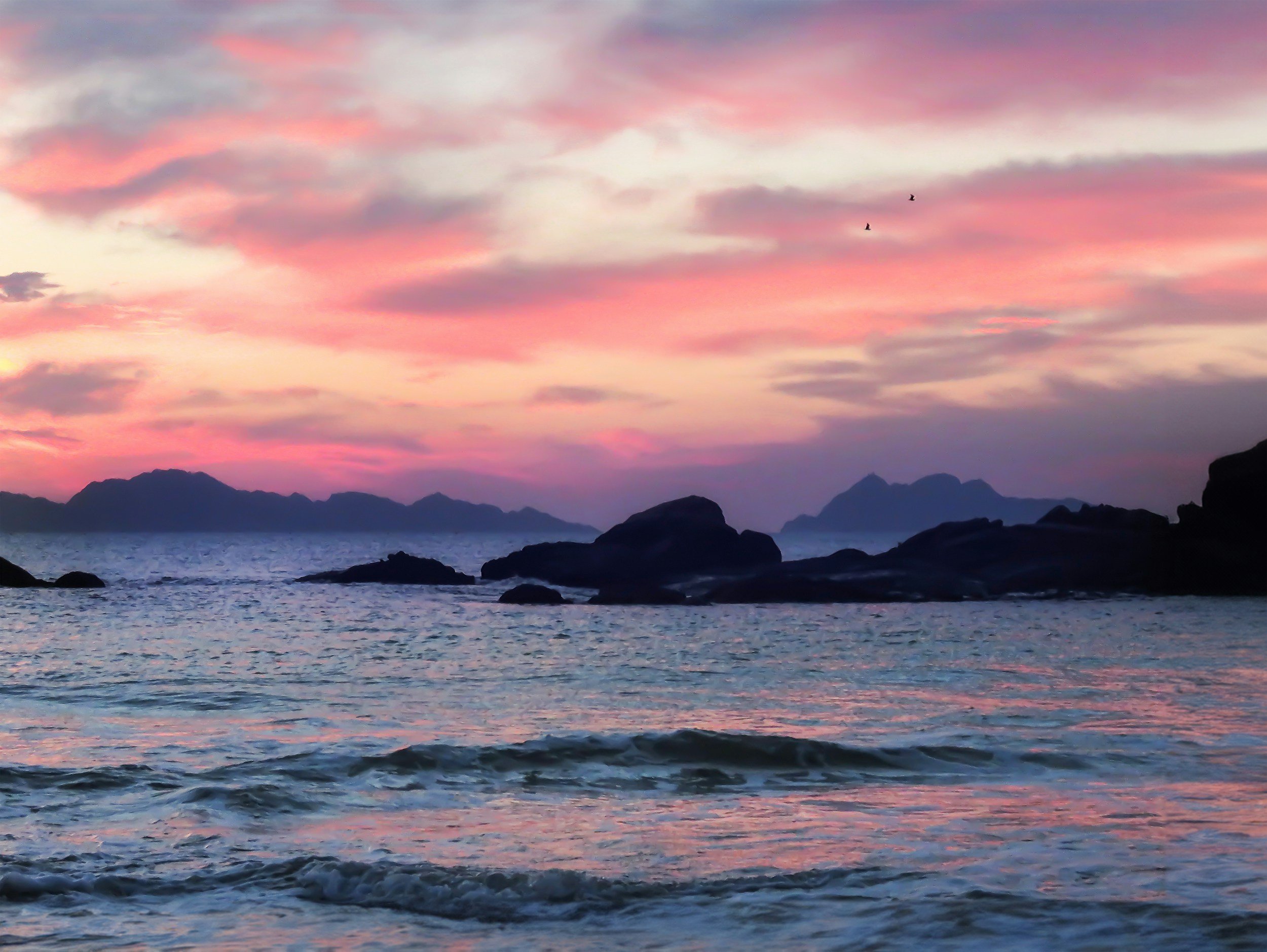 Sunset over the ocean with pink, purple, and orange clouds, silhouetted rocks in the water, distant mountains, and three birds flying in the sky.