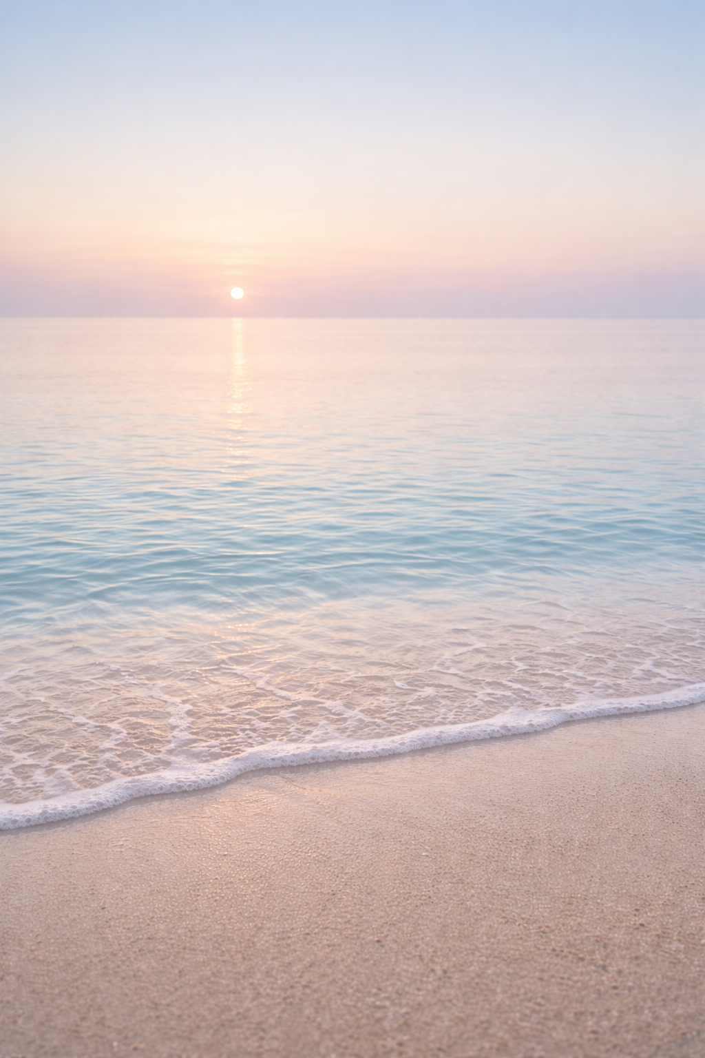 Sunset over calm ocean with pink and blue hues, gentle waves lapping sandy beach in foreground.