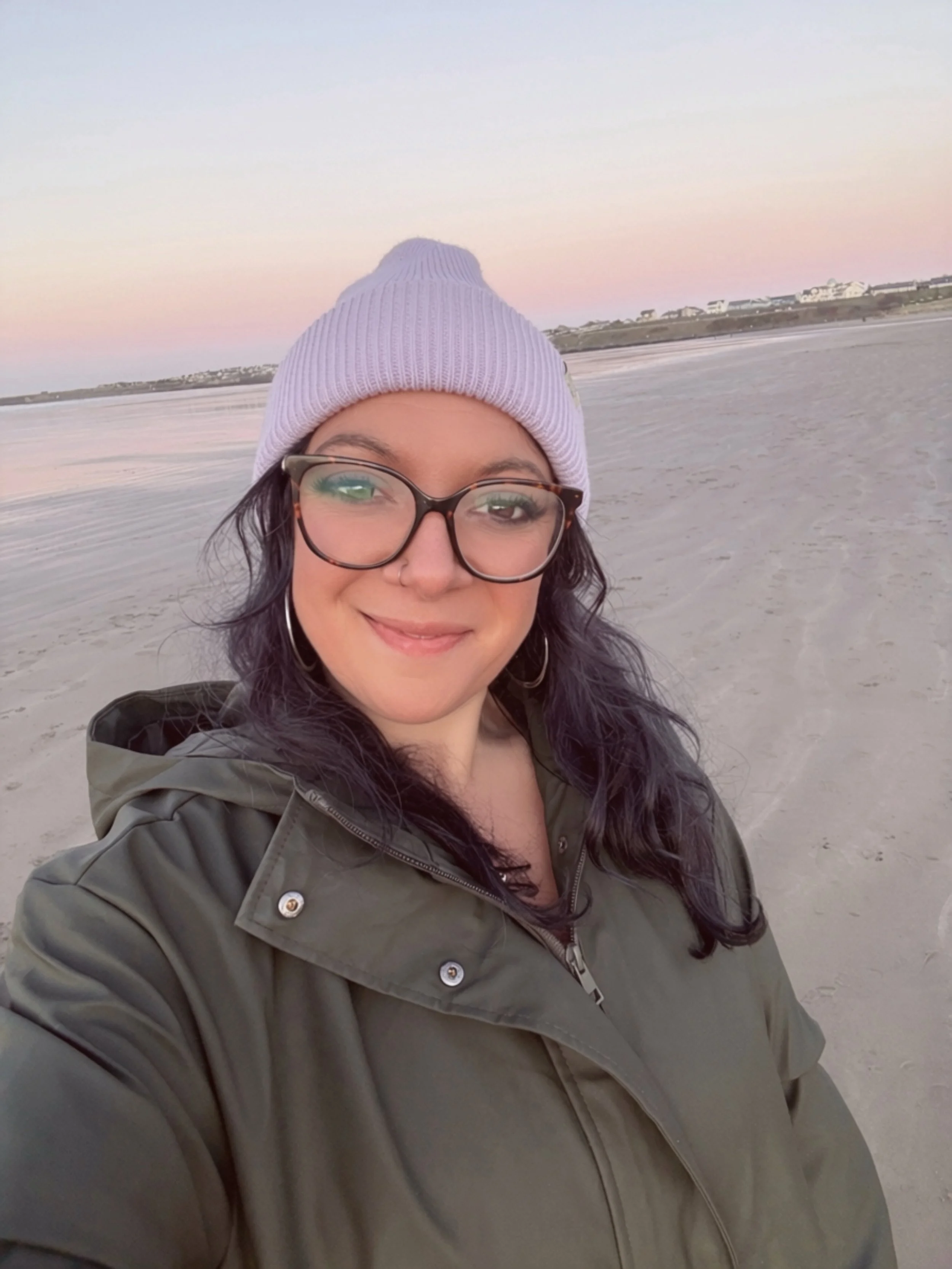 A woman taking a selfie on a beach at sunset, wearing glasses, a purple knit beanie, and a green jacket.
