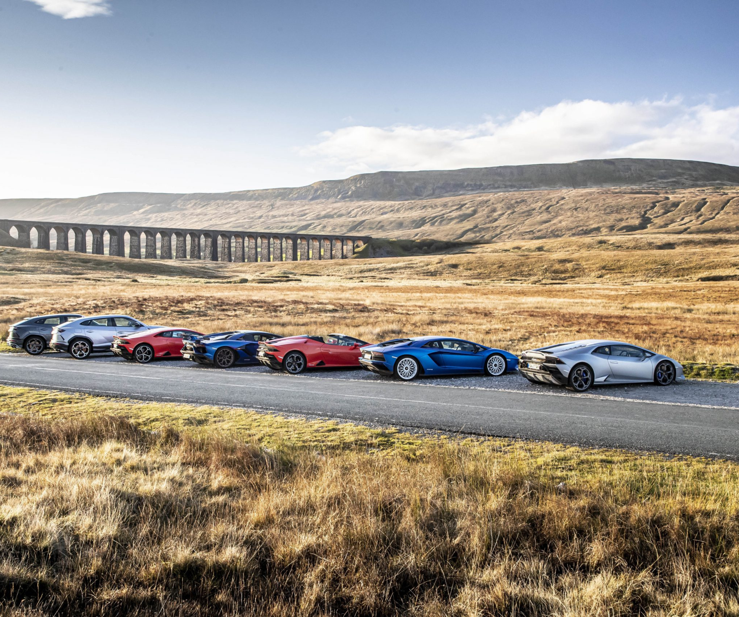 Six luxury sports cars parked on a rural road with a viaduct and rolling hills in the background.