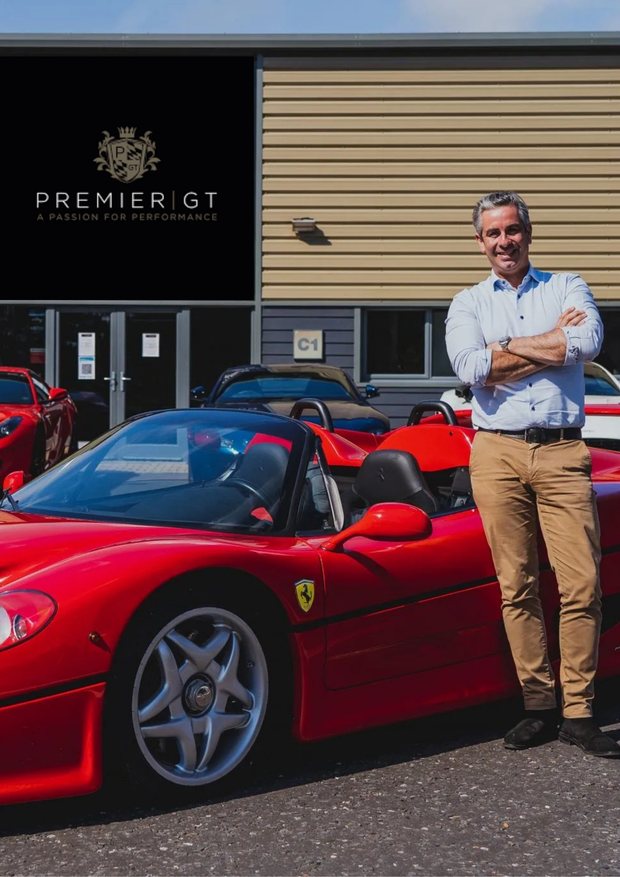 A man standing confidently with arms crossed beside a red Ferrari convertible sports car, in front of a building with a sign that reads "Premiere GT, A Passion for Performance," and other cars parked nearby.