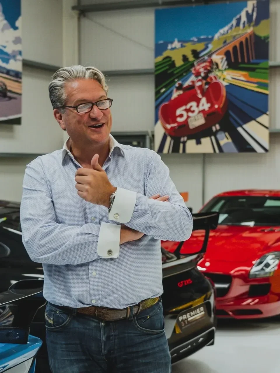 A middle-aged man with gray hair and glasses, smiling and giving a thumbs-up, standing in a room with luxury cars and racing-themed artwork on the wall.
