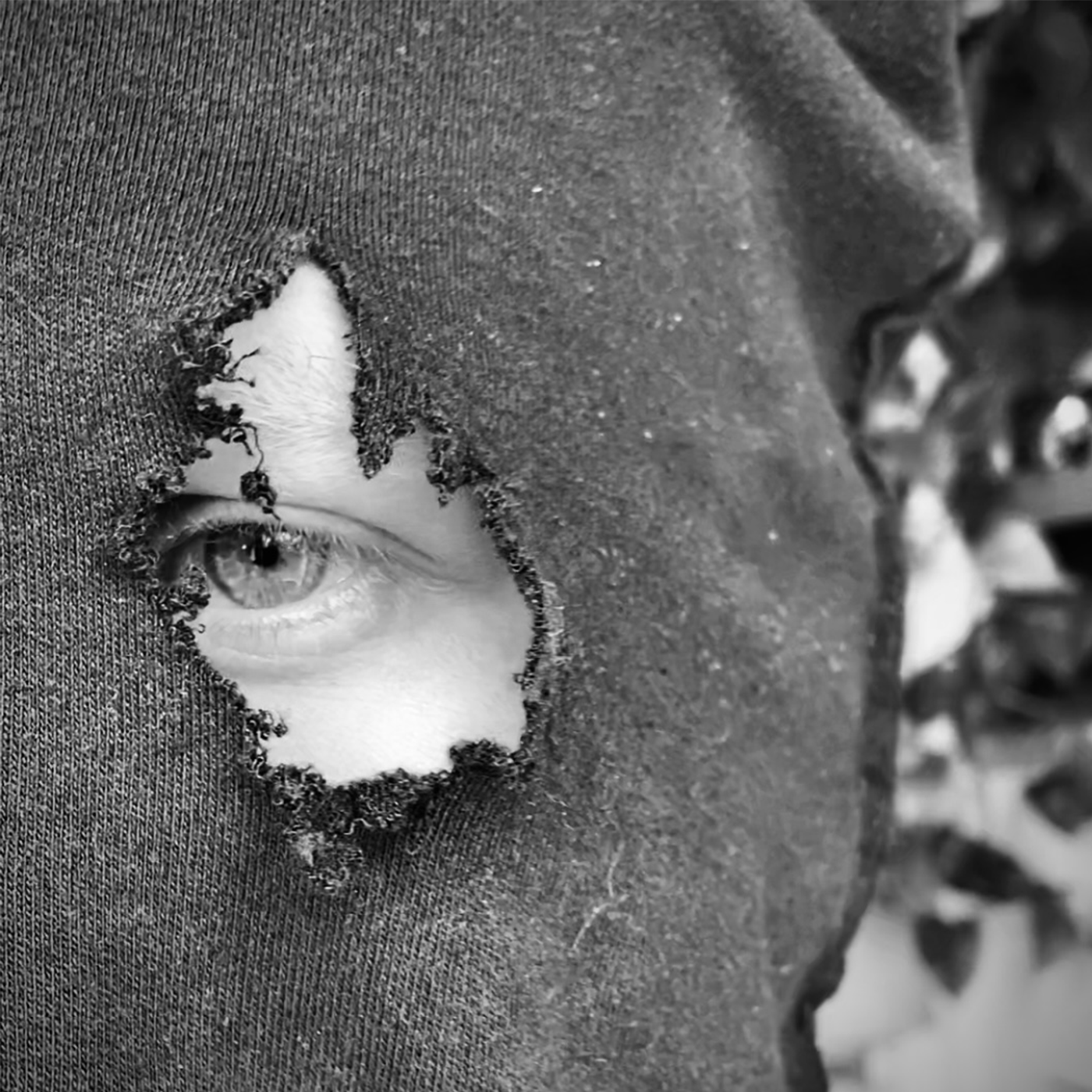 A person's eye peeking through a hole torn in a fabric or paper, with plants or foliage in the background.