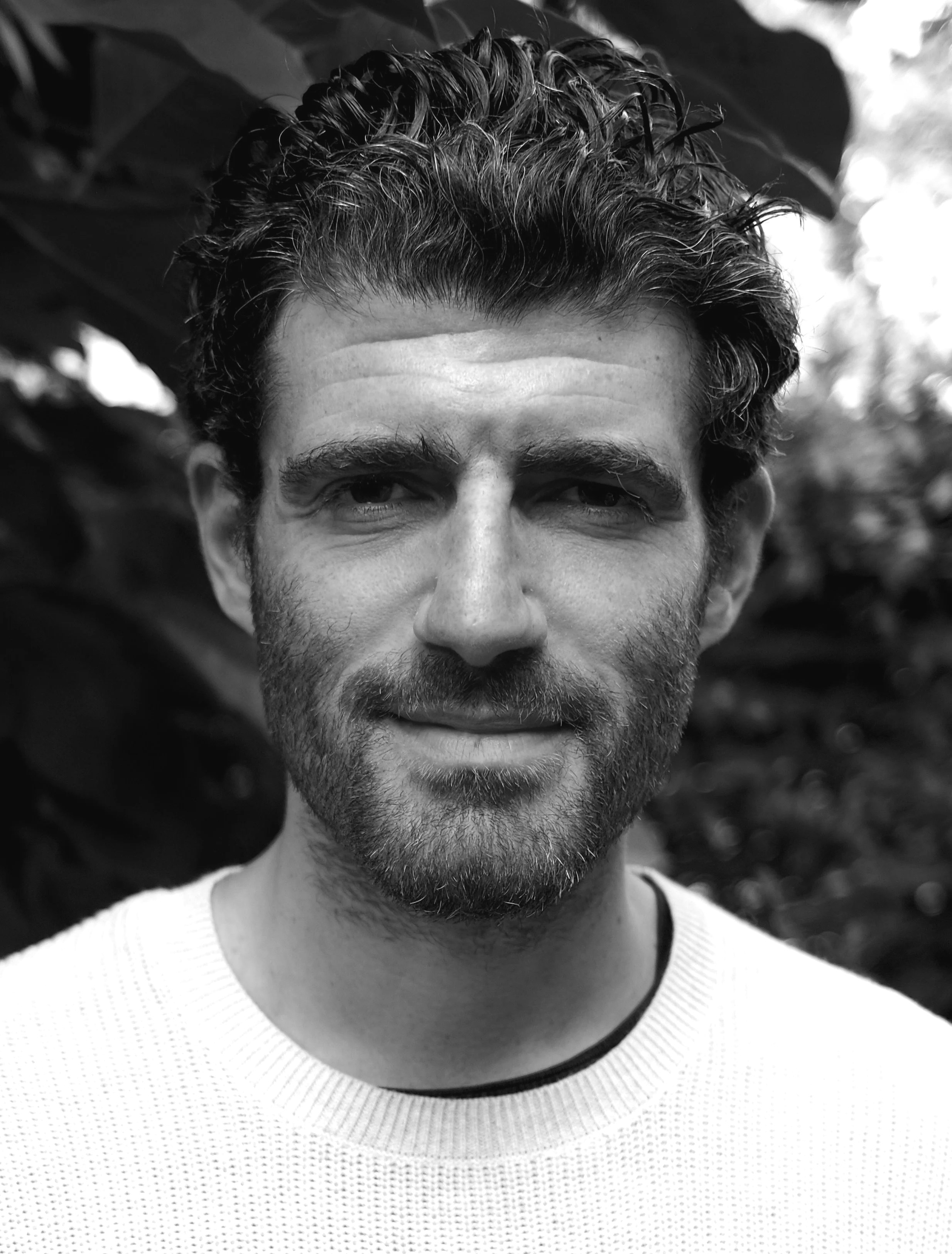 Black and white close-up portrait of a man with dark, wavy hair, a beard, and wearing a light-colored sweater, standing outdoors with blurred foliage in the background.