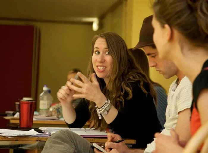 A woman with long brown hair making a funny face, sitting at a table with others, holding a smartphone, in an indoor setting.