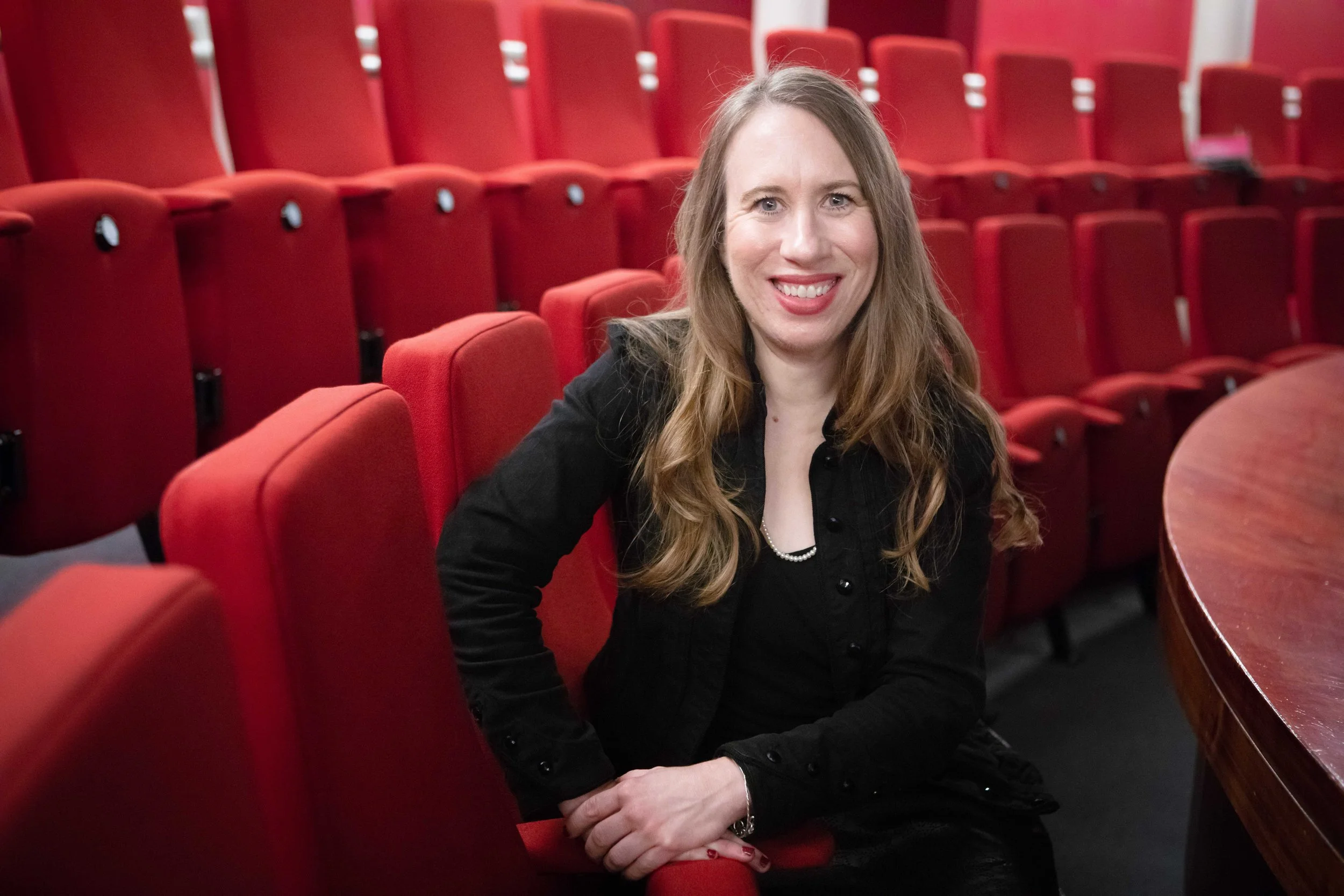 A smiling woman with long wavy hair sitting in red auditorium chairs, wearing a black jacket and pearl necklace, next to a curved wooden table.