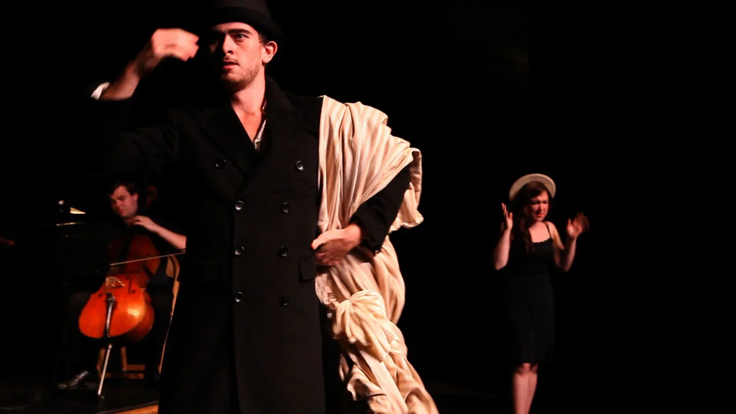 The image shows a theatrical performance with three actors on stage. The male actor in the foreground is dressed in a black coat and a beige shawl, gesturing with his hand. In the background, a female actor in black clothing and a white hat appears t