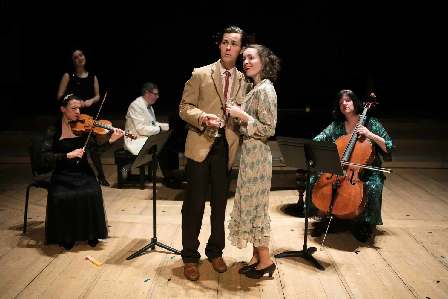 A group of five musicians performing on stage, including two singers in the center, with a violinist, pianist, and cellist surrounding them.