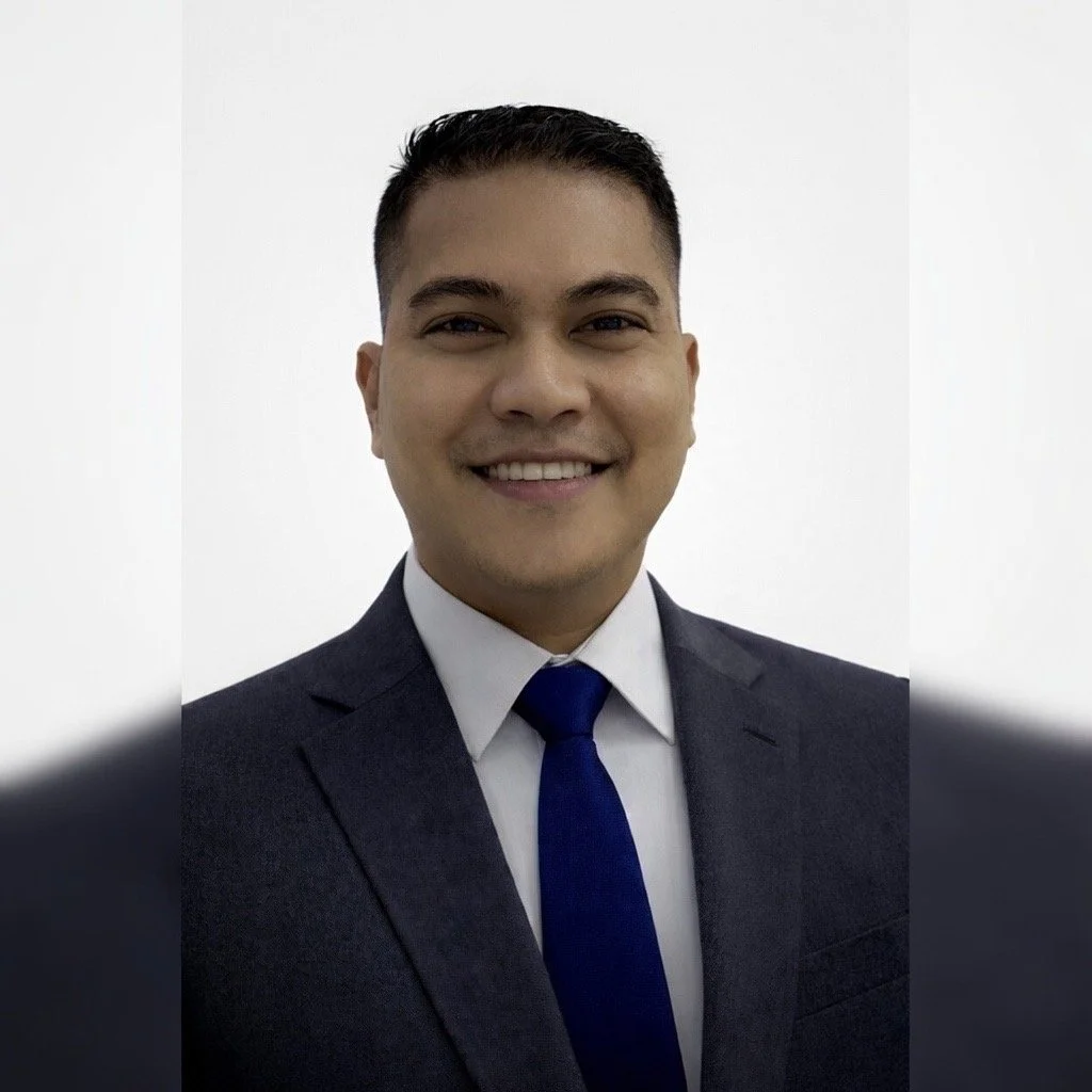 A young man with short dark hair smiling, wearing a gray suit, white shirt, and blue tie against a plain gray background.