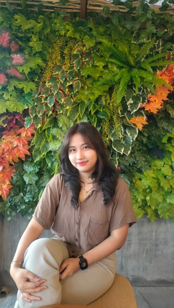 A young woman with dark hair, wearing a brown shirt and beige pants, sitting on a stool in front of a vibrant, colorful wall of assorted green and autumn-colored foliage.