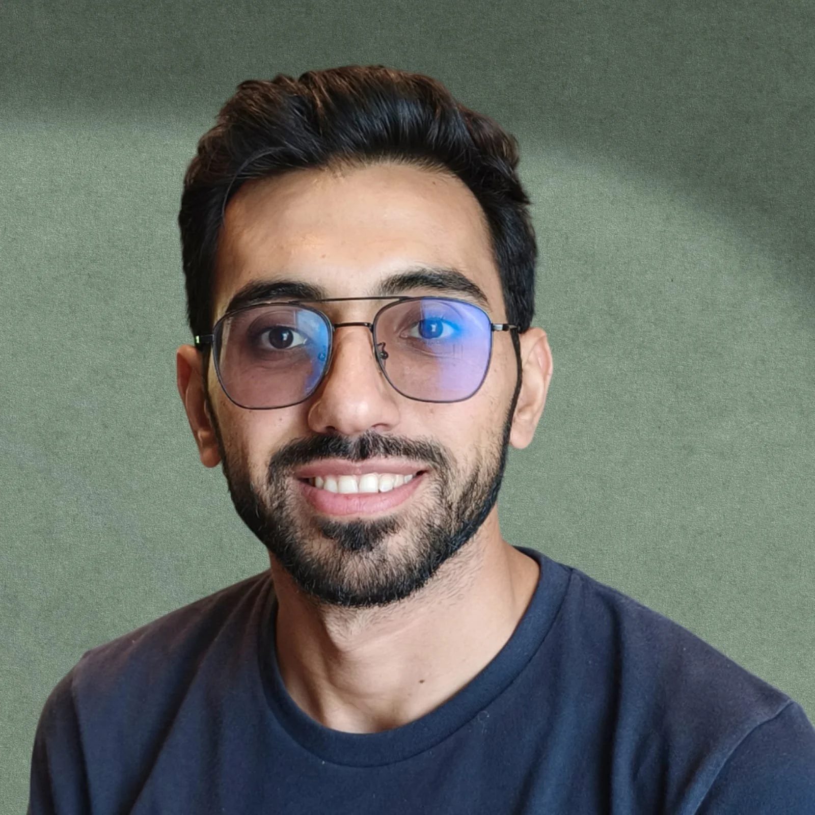 Portrait of a man with short dark hair, glasses reflecting blue light, a beard, and a smile, wearing a dark t-shirt, against a green background.