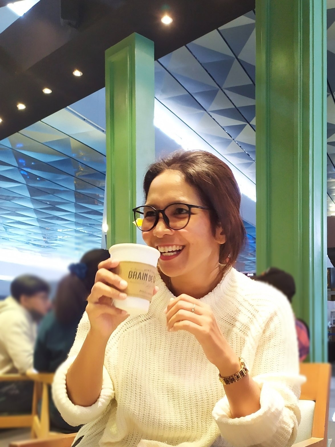 A woman with glasses smiling and holding a coffee cup in a busy café with green pillars and a patterned ceiling.