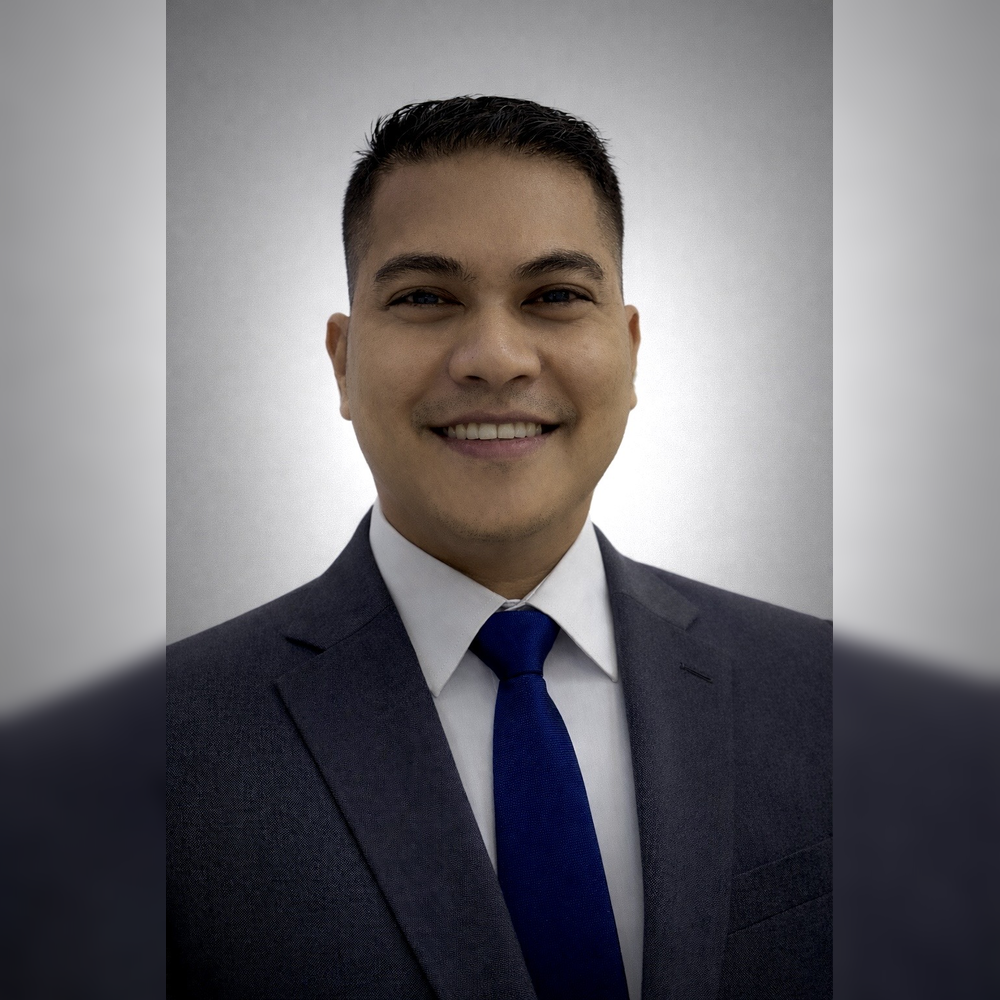 A professional young man smiling, wearing a suit, white shirt, and blue tie, with a plain gray background.