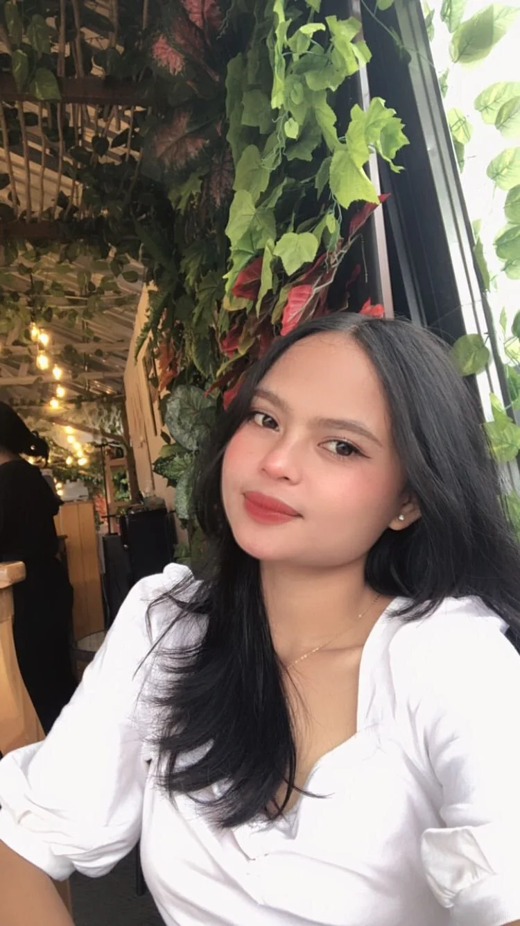 A young woman with long black hair wearing a white top, sitting indoors near a window decorated with green plants, with a warm and cozy ambiance in the background.