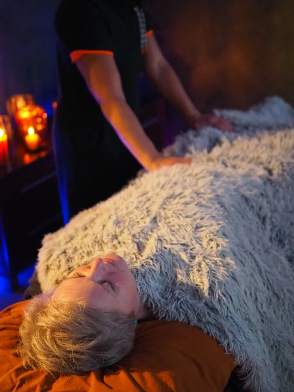 Nuori nainen makaa selällään pehmeällä harmaatäpläisellä peitteellä ja levähtää silmät suljettuina, ympärillä on hämyinen valaistus, kynttilöitä ja henkilöitä seisomassa hänen päällään.