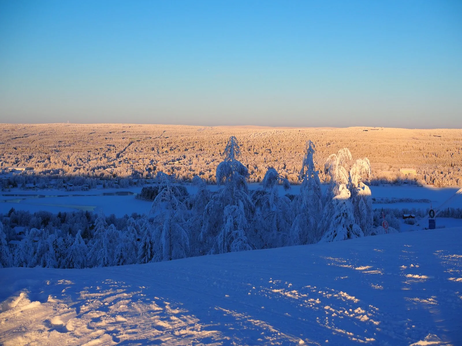 Luminen talvimaisema auringonlaskun aikaan, jossa on lumisia puita ja taustalla kaupunki tai kylä vuoristojen keskellä.