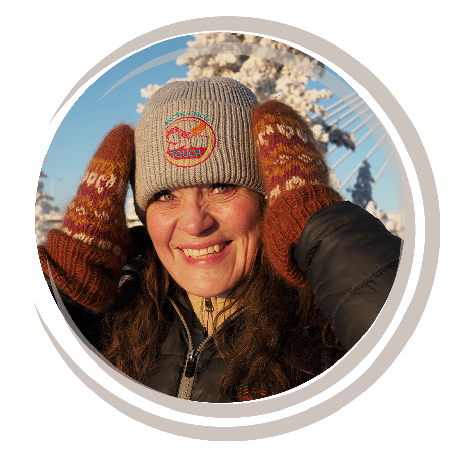 Nainen hymyilee talvisella säällä, pukeutuneena pipoihin ja lapasiin, taustalla luminen maisema ja puut.