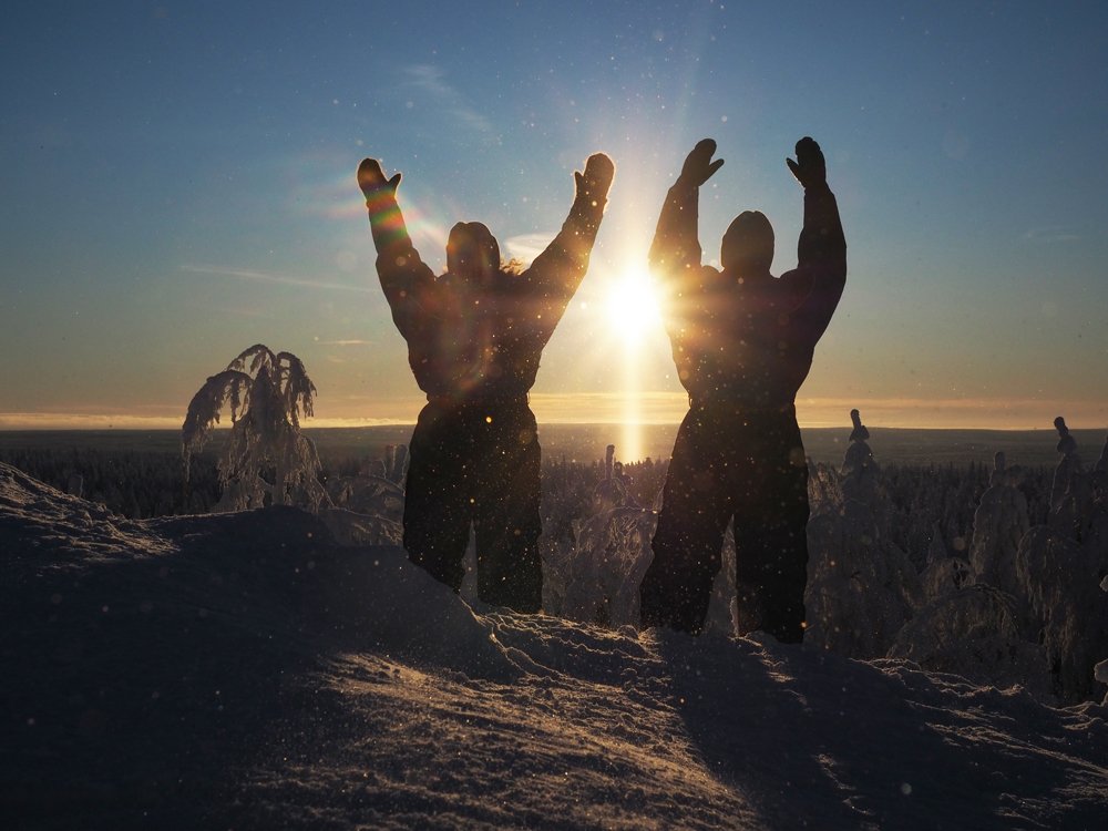 Kaksi ihmistä seisoo lumisessa maisemassa auringonlaskun aikaan, heidän kädet kohoavat ilmaan ja heidän taustalleen tulee auringon säteet.