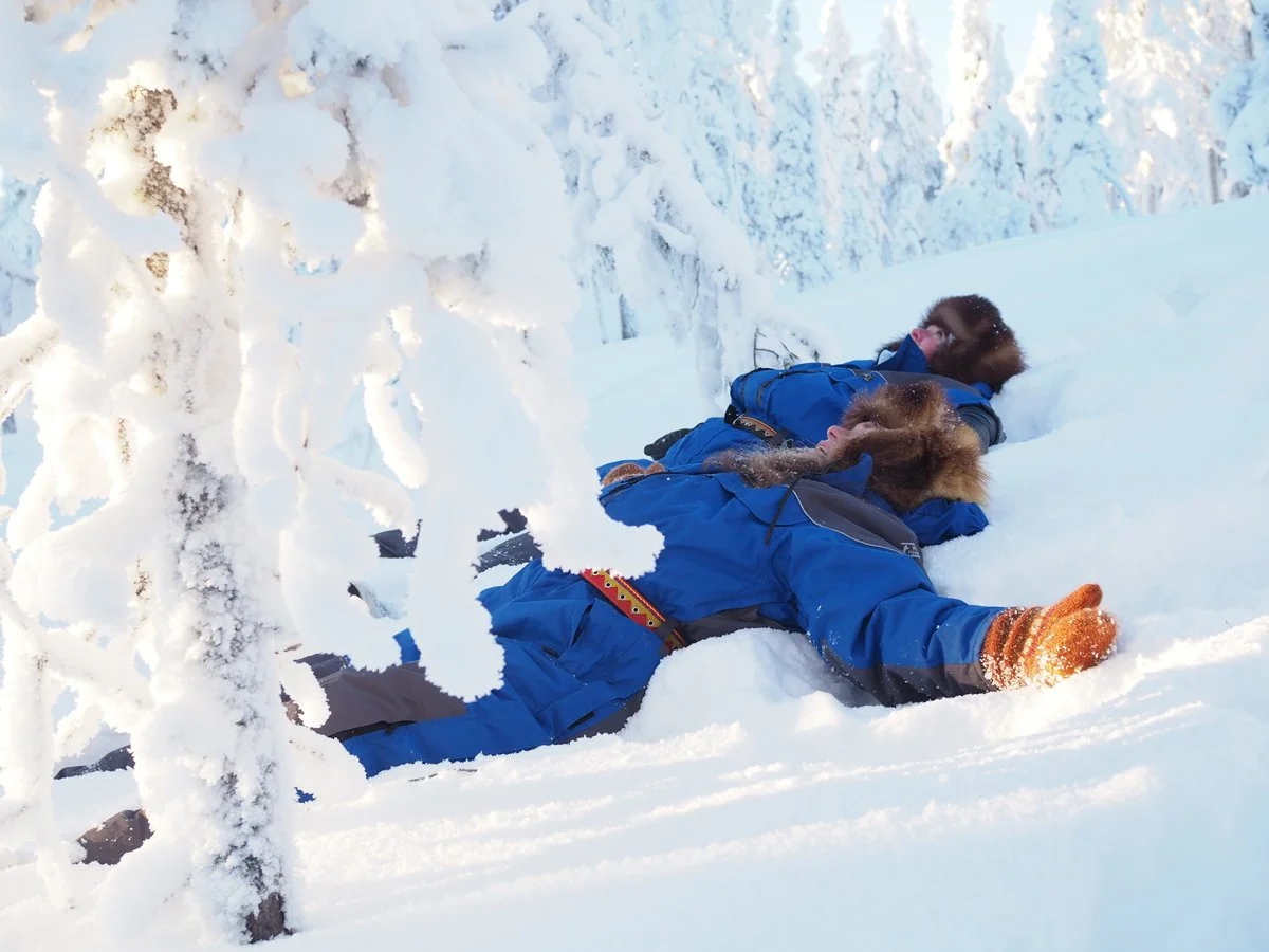 Kaksi henkilöä makaa lumessa talvisessa maisemassa, ympärillään lumisia puita, molemmat pukeutuneet sinisiin talvivaatteisiin ja pipoihin.