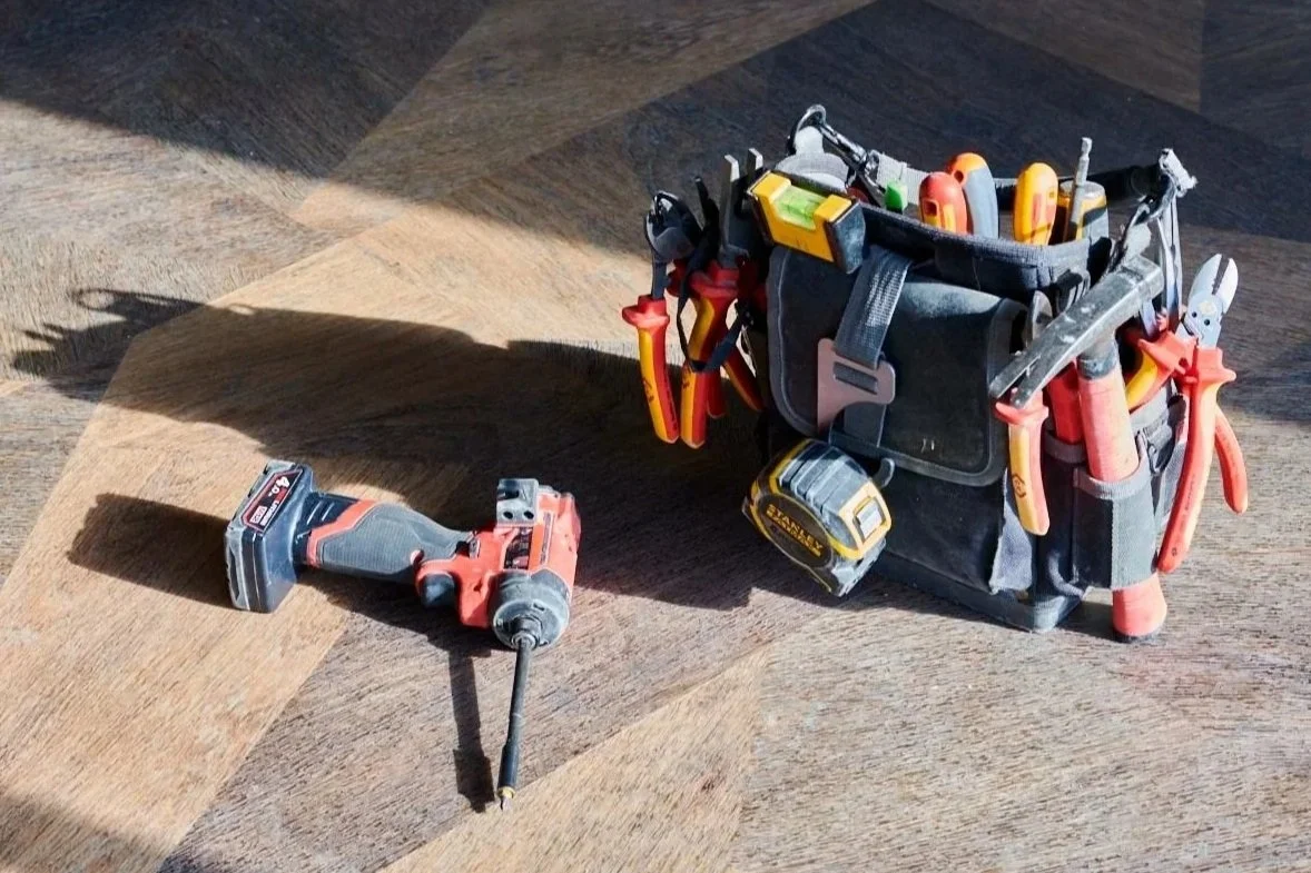 A cordless drill and a tool bag filled with various hand tools and power tools on a wooden surface.