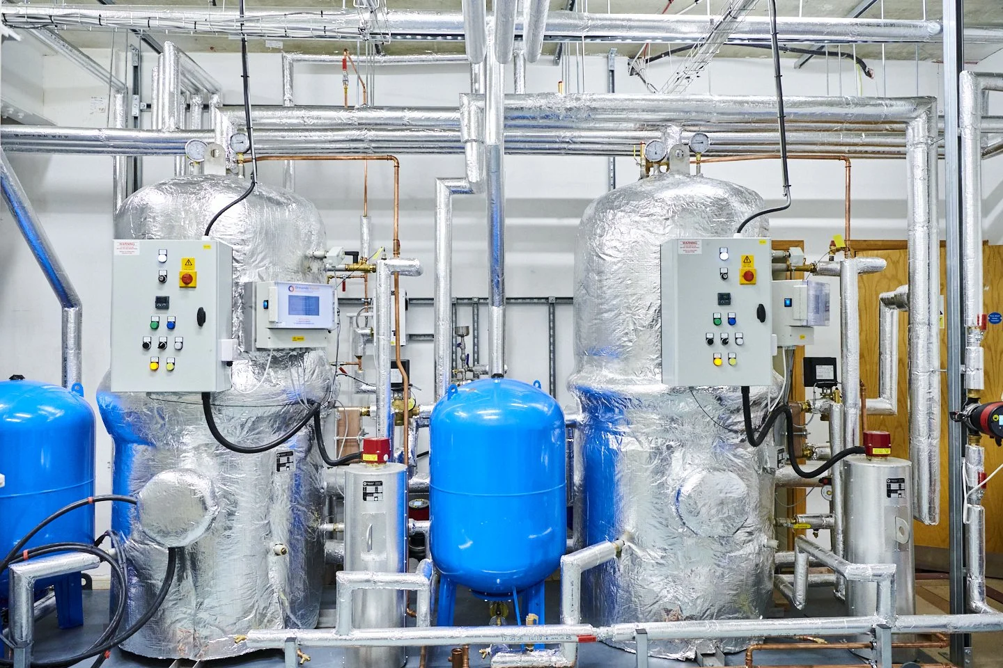 Industrial equipment with two large silver insulated tanks, blue pressure tanks, and numerous pipes and control panels in a mechanical room.