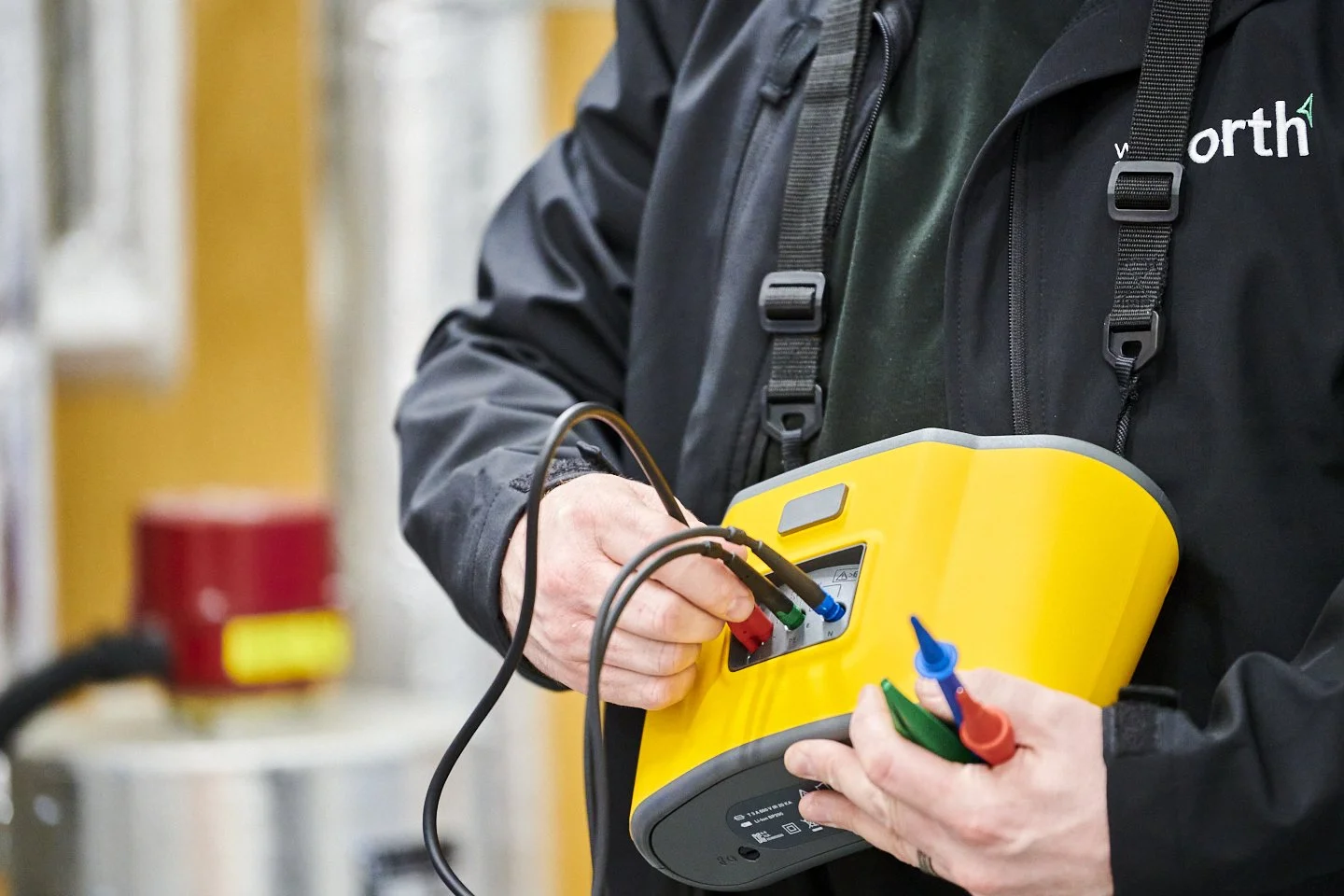 A person wearing a black jacket and a black backpack is holding a yellow electrical testing device with probes. They are connecting the probes to the device, which has a display and multiple colored ports. The background is an industrial setting with blurred equipment.