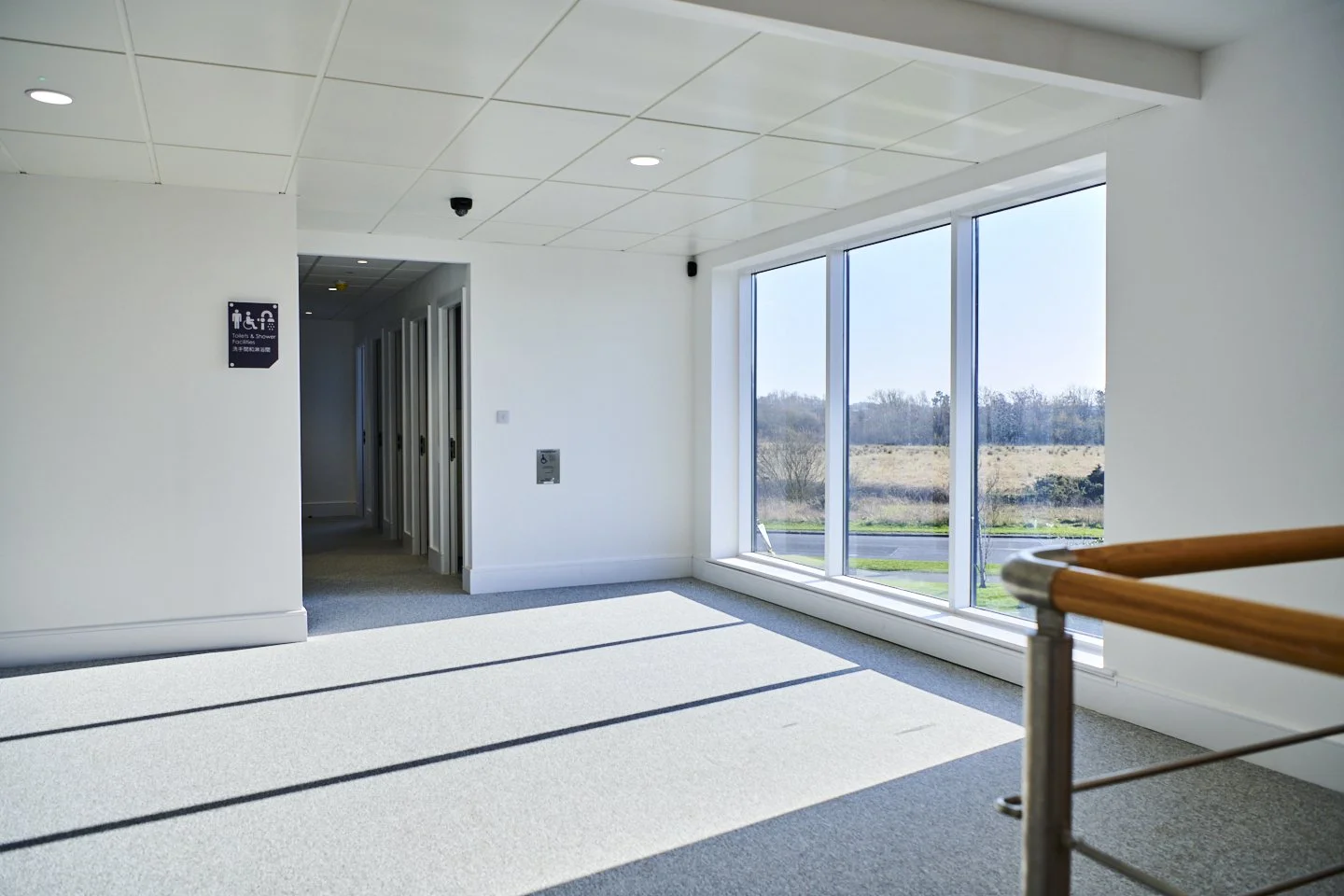 Bright indoor space with large windows revealing an outdoor landscape, white walls, and a wooden railing.