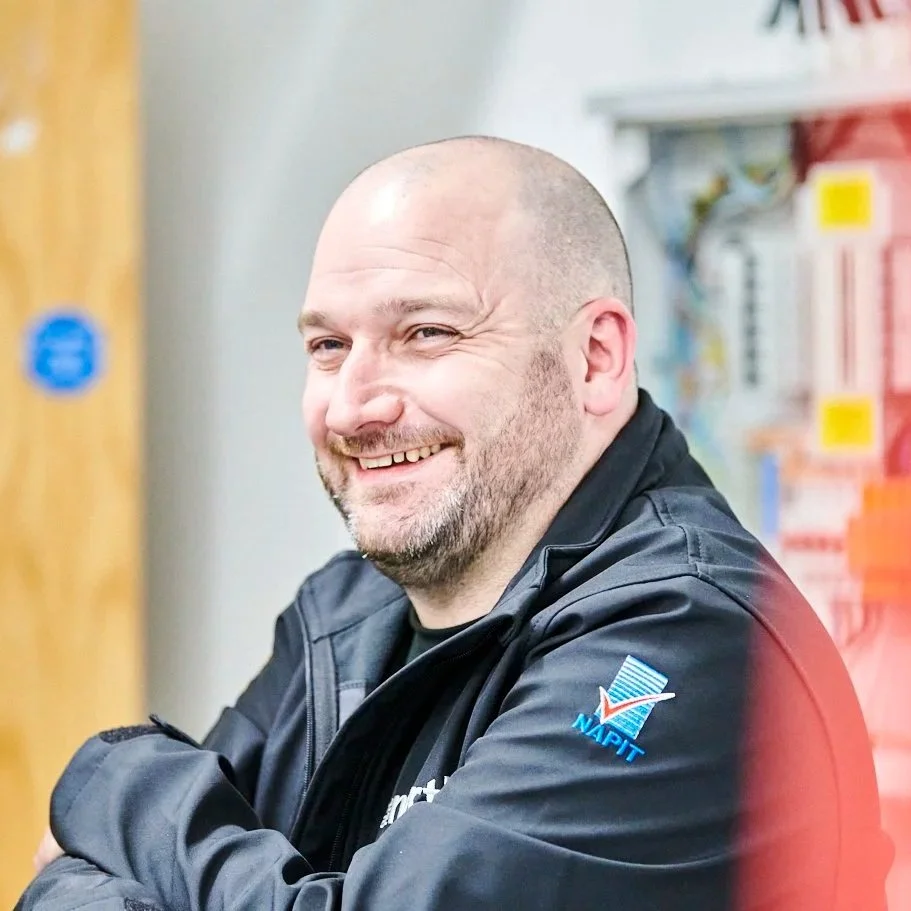 Smiling man with a beard and short hair sitting indoors, wearing a black jacket with NAPIT logo, against a background of store shelves and a wooden door.