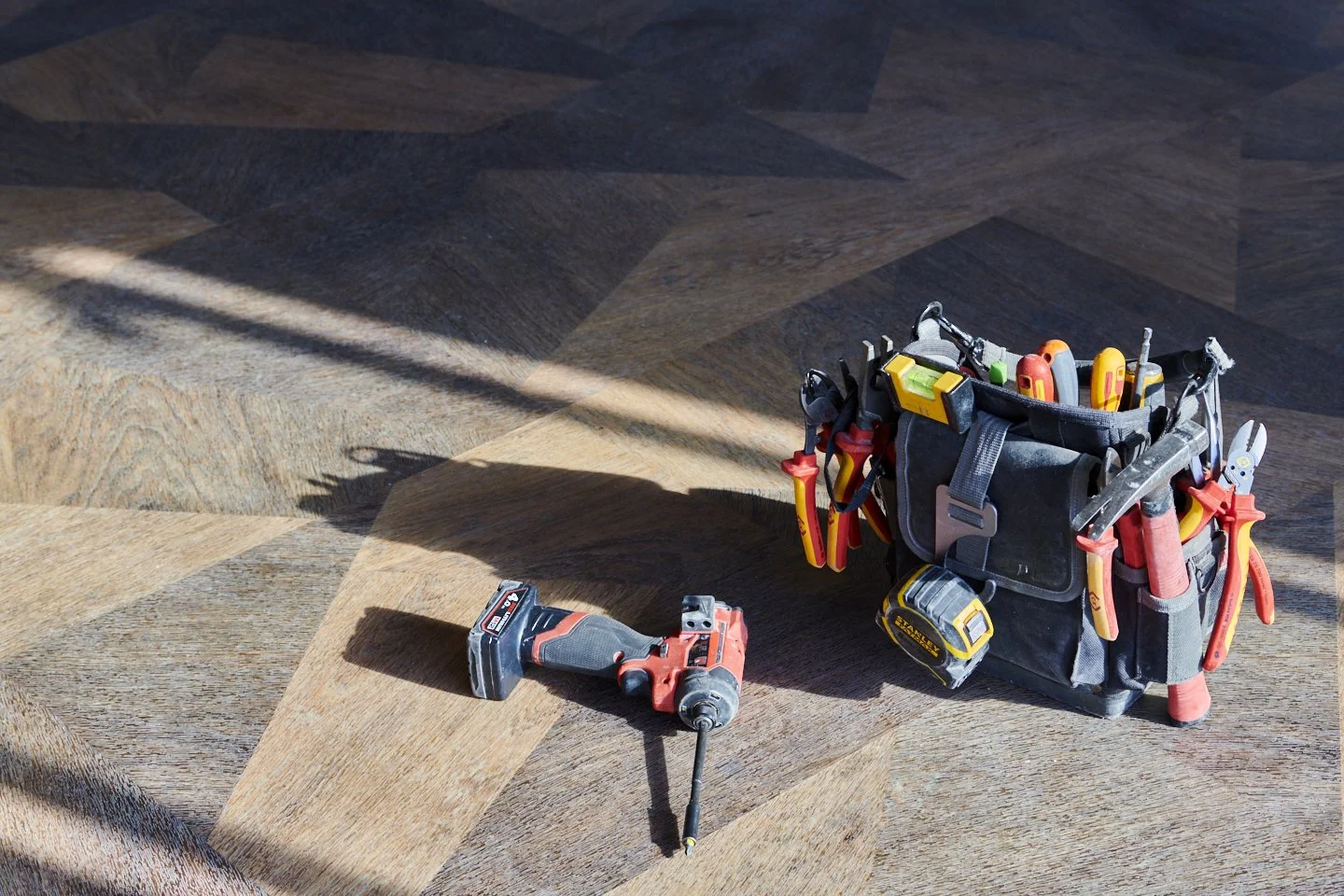 A cordless power drill and a tool belt filled with various hand tools are placed on a hardwood floor illuminated by natural light.